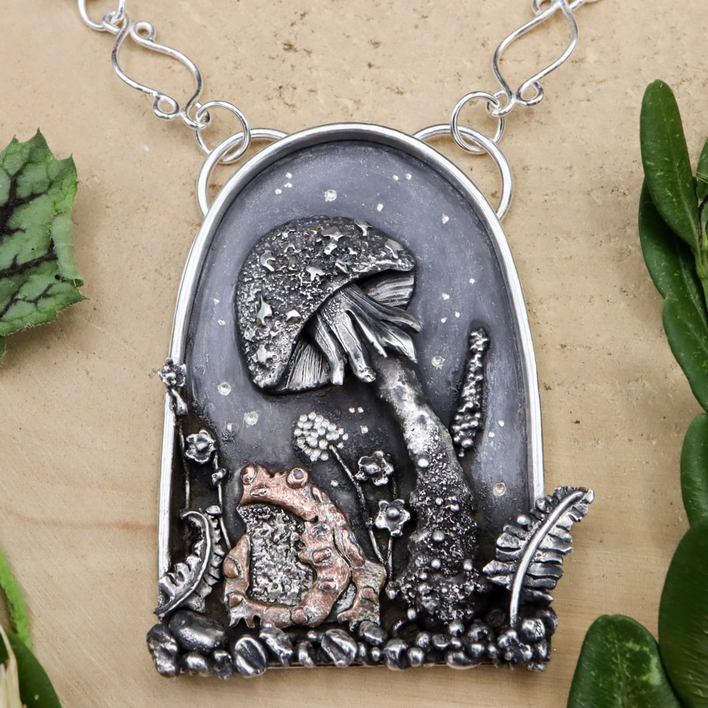A sterling silver and copper toad pendant. The pendant features the toad at the bottom of it sitting under a mushroom toadstool. There are little silver flowers and ferns around them. The necklace is shown on a piece of light tan wood.