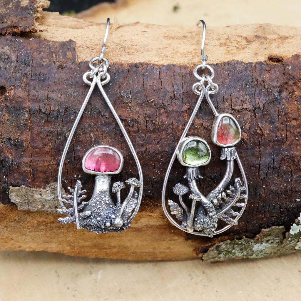 A pair of mismatched watermelon tourmaline mushroom dangle earrings are shown on a piece of darn brown wood. Each earring has a gemstone capped mushroom as the focal point.