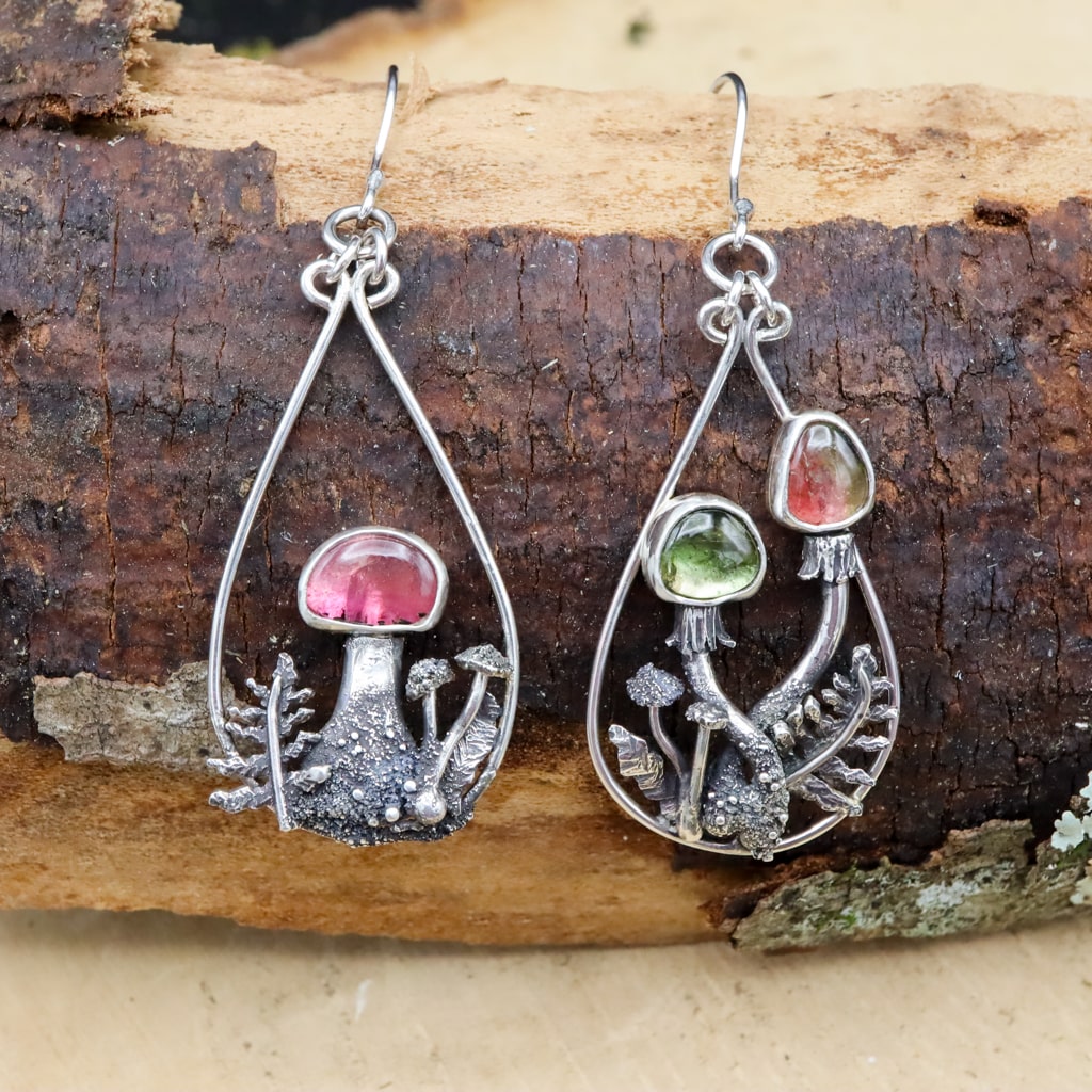 A pair of mismatched watermelon tourmaline mushroom dangle earrings are shown on a piece of darn brown wood. Each earring has a gemstone capped mushroom as the focal point. 