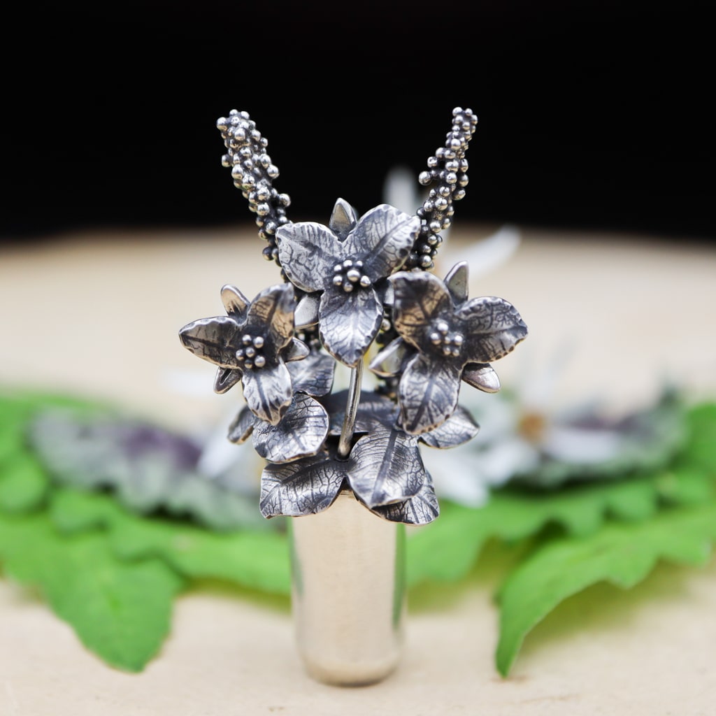A sterling silver necklace featuring a tiny silver vase holding 3 hand carved trillium flower. The pendant is about 2.25 inches tall and shown with greenery in the background.