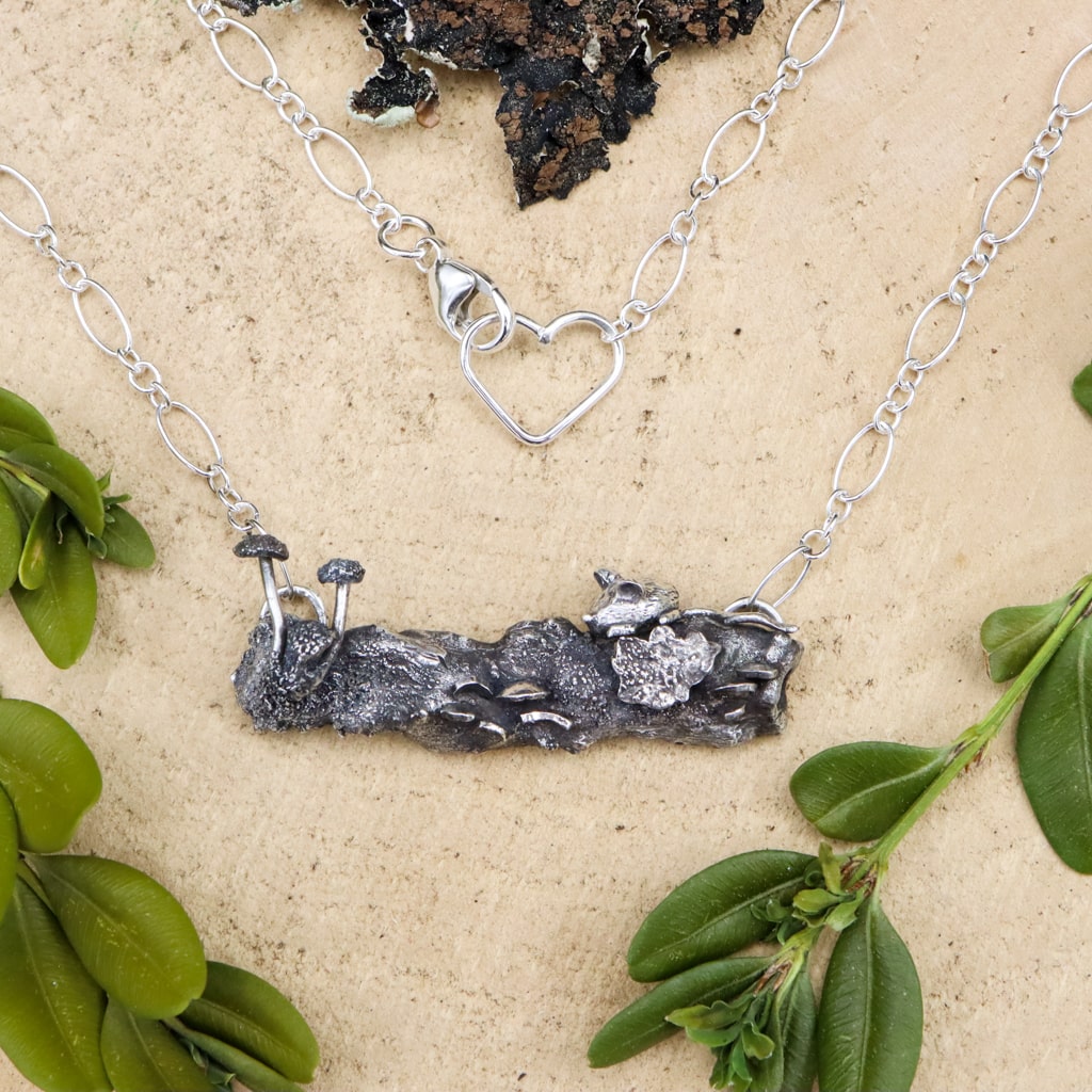 A handmade sterling silver necklace that resemble a forest log. On the silver log there is a tiny silver mouse sitting above some lichen, turkey tail mushrooms, and some button mushrooms. The necklace is shown on a piece of light tan wood and surrounded by greenery.