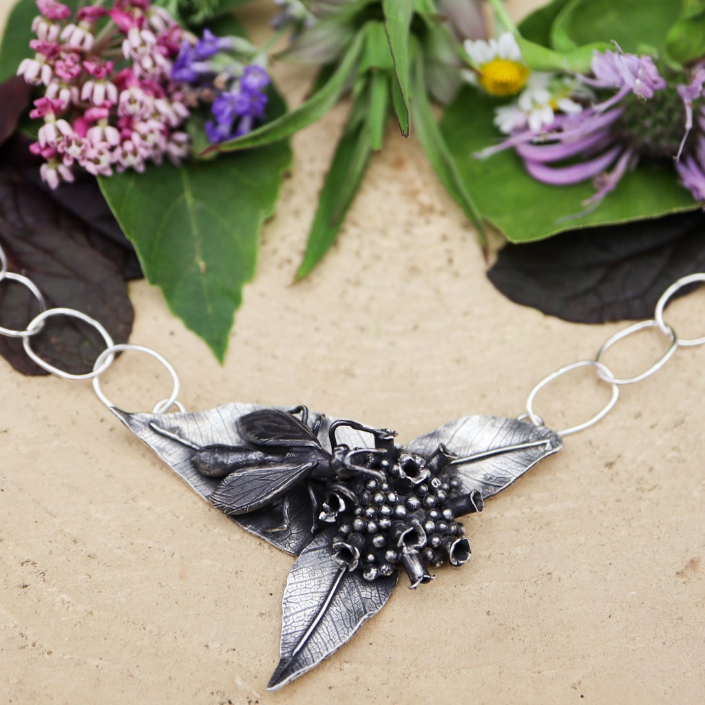 A native great black wasp was carved from sterling silver and sits upon a silver mountain mint flower. It is a necklace with a loop chain and shown with real flowers in the background on a piece of tan wood.