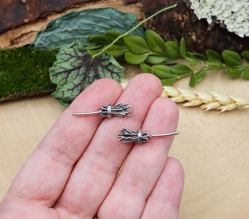 A hand is shown holding a pair of sterling silver witches broom earrings for size. They are about 1 inch wide. 