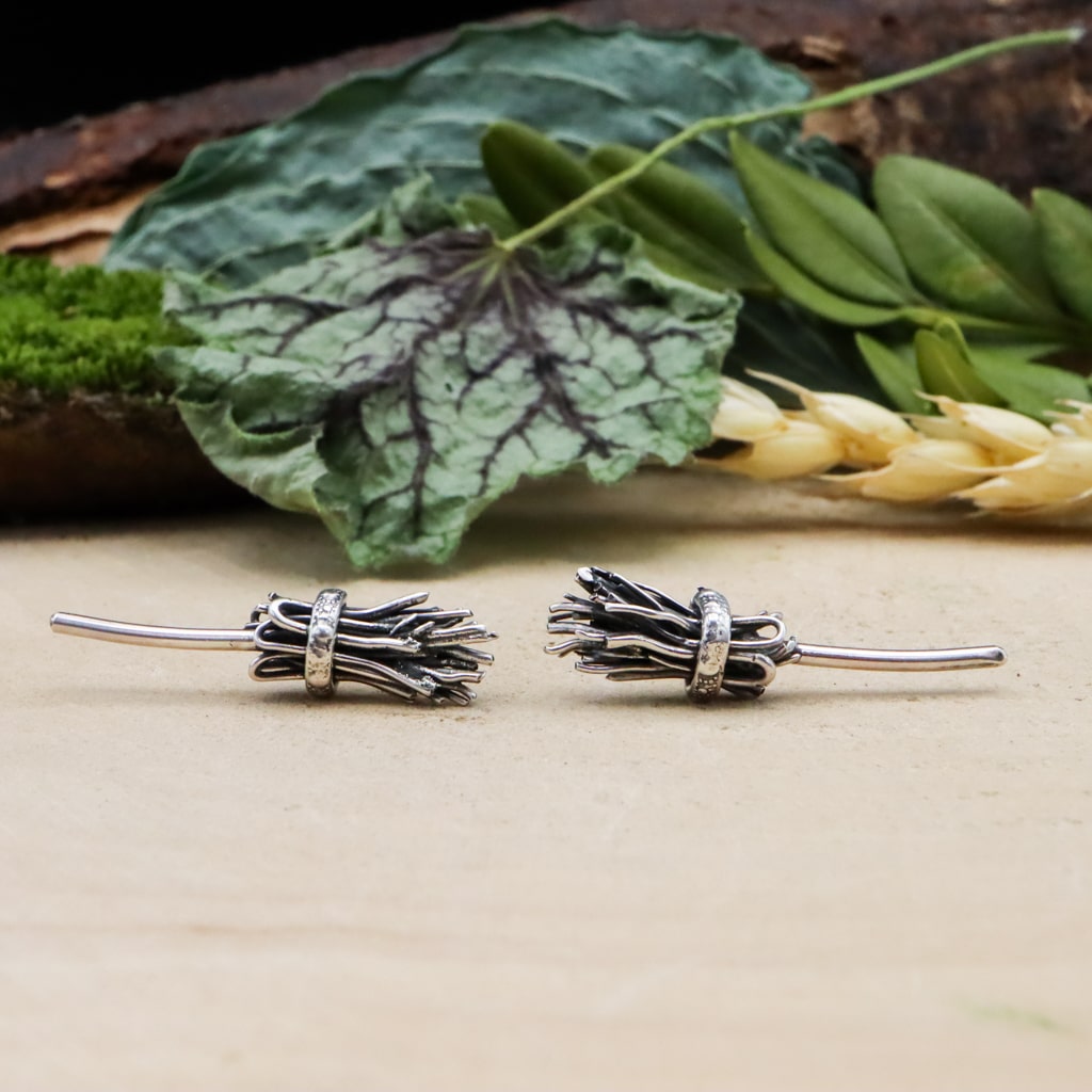 A pair of handmade sterling silver witches broom earring studs. They are about 1 inch wide and shown on a light tan piece of wood. 