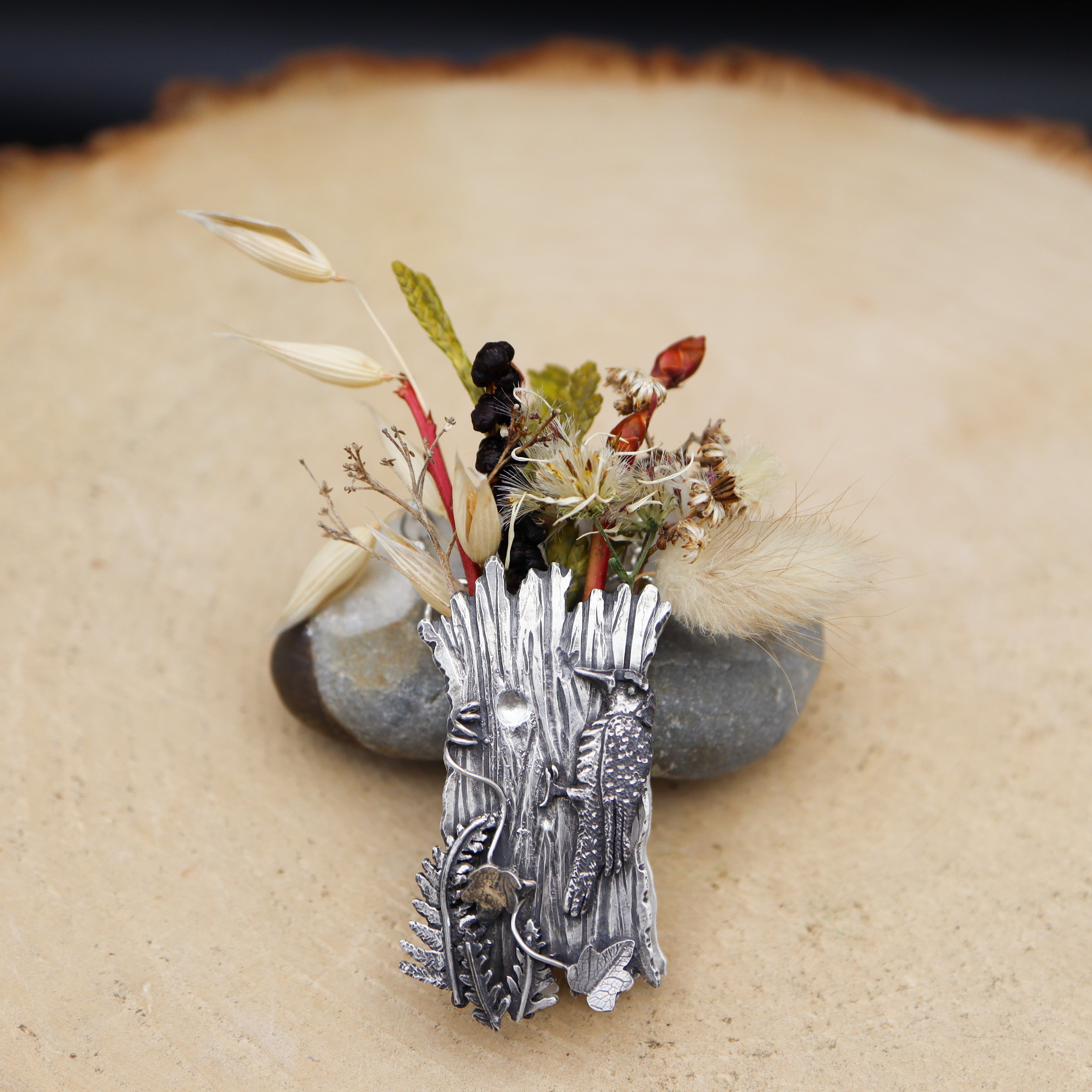 A sterling silver woodpecker vase necklace with some ferns and ivy vine growing on the front of it. The vase is about 2 inches tall and one of a kind. It is shown leaning on a light grey stone on a tan piece of wood. There are some brightly colored twigs and plants sitting inside the little vase necklace.