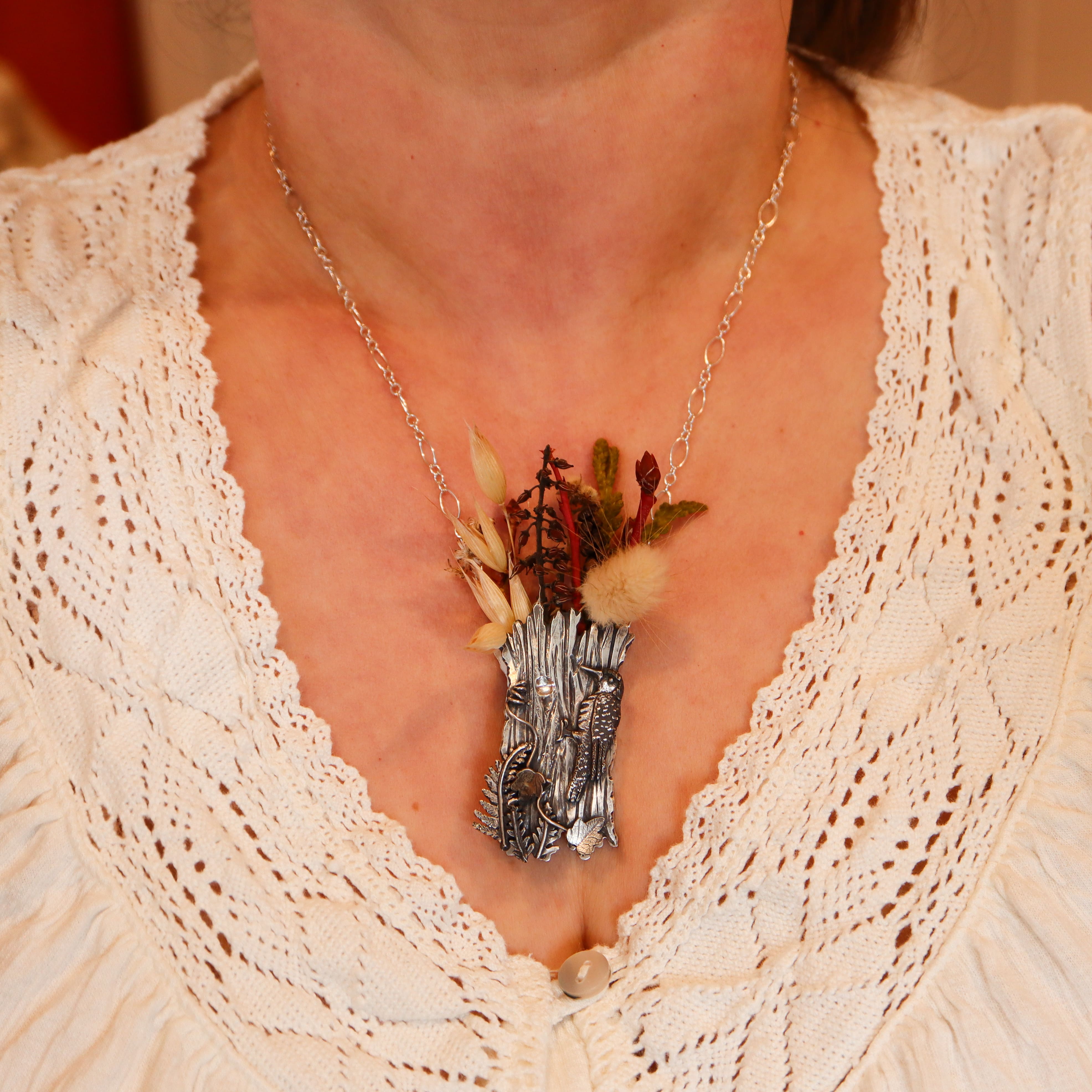 A woman is shown wearing the vase necklace with flowers and twigs in it. She is wearing a white lacy v neck blouse.