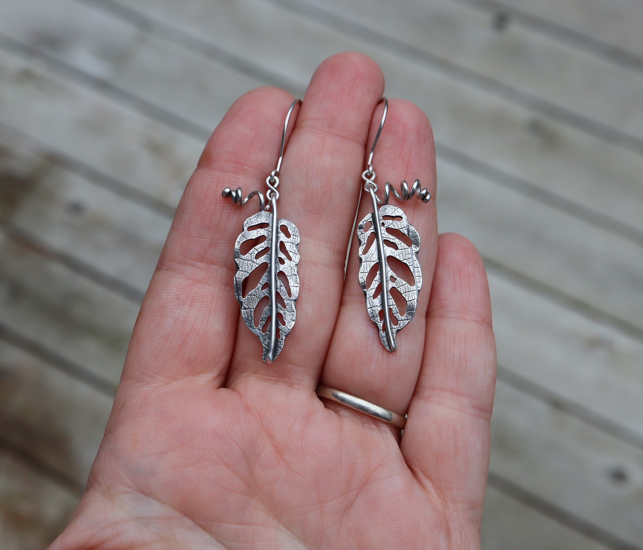 Sterling silver handmade monstera adansonii dangle earrings made by The Striped Cat Metalworks. They are pictured in a hand to show size.