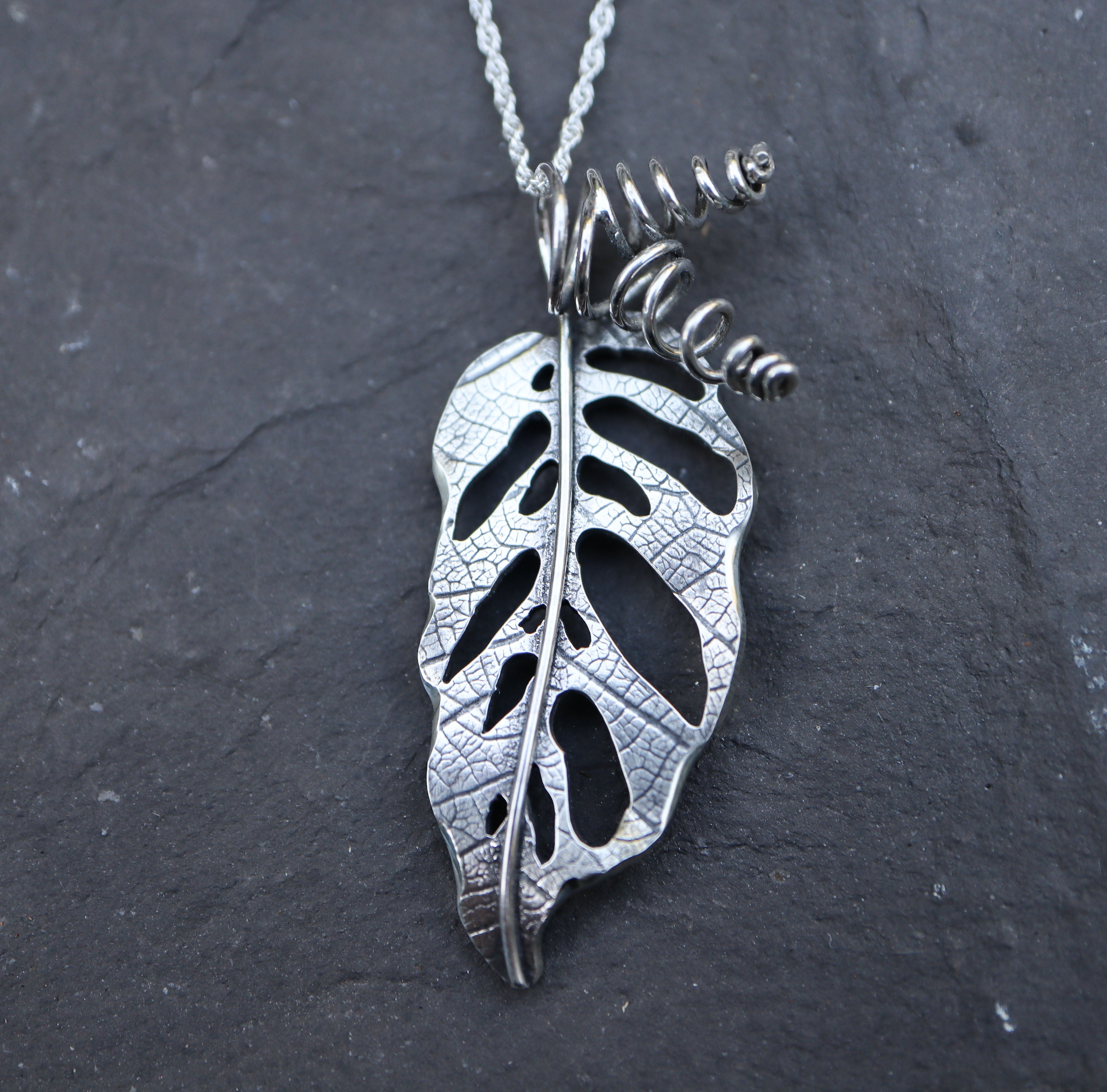 A handmade sterling silver monstera adansonii necklace made by The Striped Cat Metalworks. The necklace is pictured on a dark grey piece of slate.