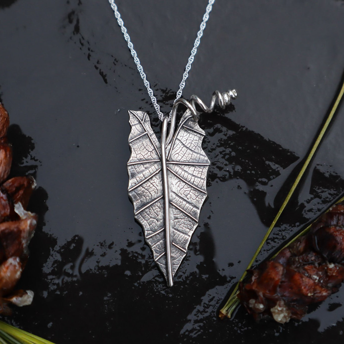A handmade sterling silver alocasia houseplant leaf necklace. The leaf is about 1.25 inches tall and has a twisty stem holding it onto a necklace.