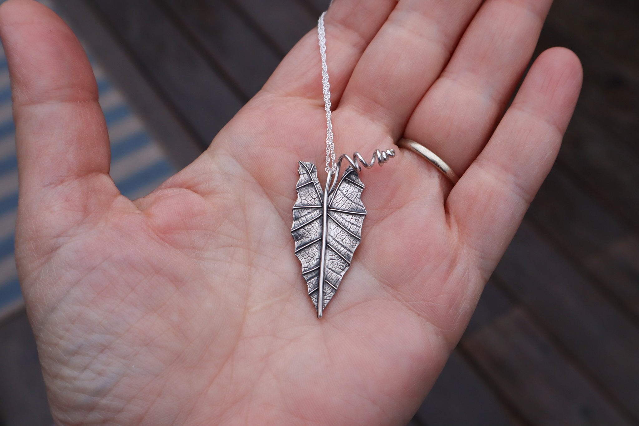 A hand is shown holding the sterling silver alocasia houseplant leaf necklace for size reference. It is about 1.25 inches tall.