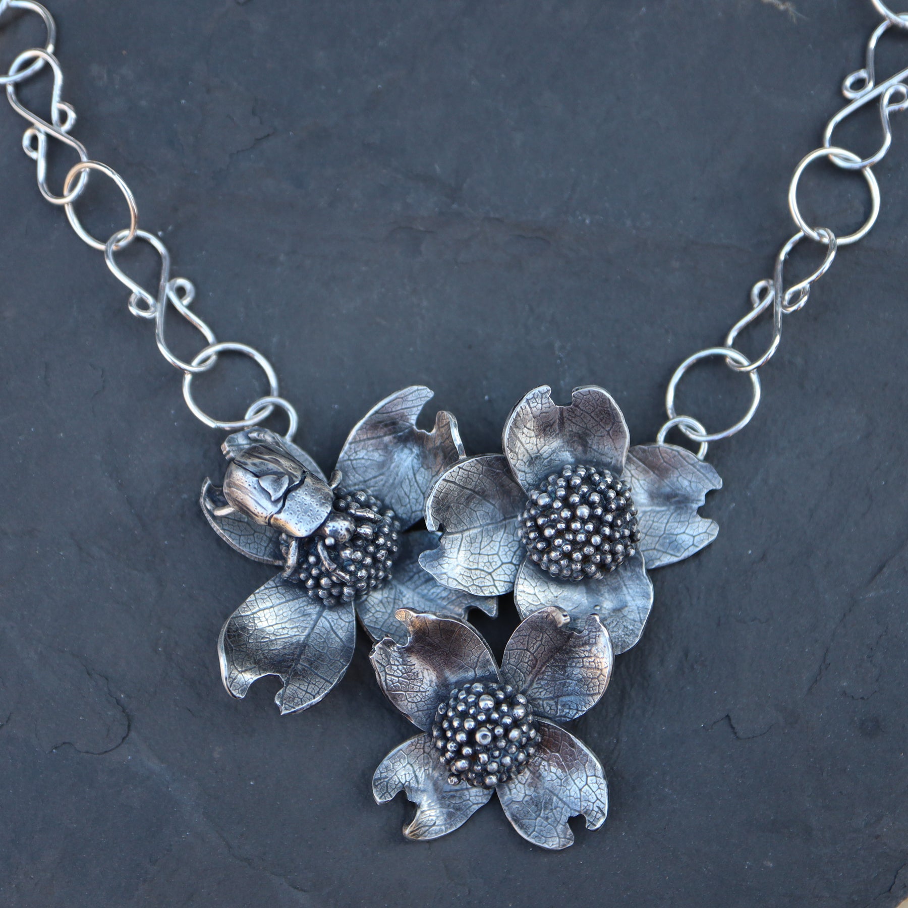 A handmade sterling silver dogwood flower and beetle necklace shown on top of a dark grey piece of slate.