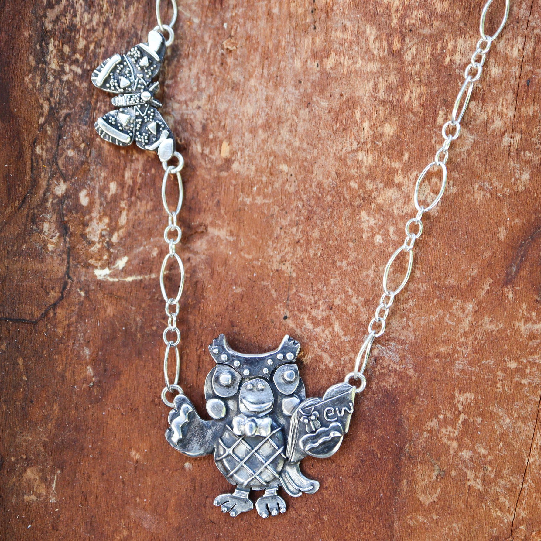 Animal Crossing Blathers necklace with an atlas moth to the left of the necklace. Blathers is holding a little book with a tiny butterfly on it and the word "ew" next to it. This necklace is made from sterling silver and shown on a dark reddish brown piece of wood.