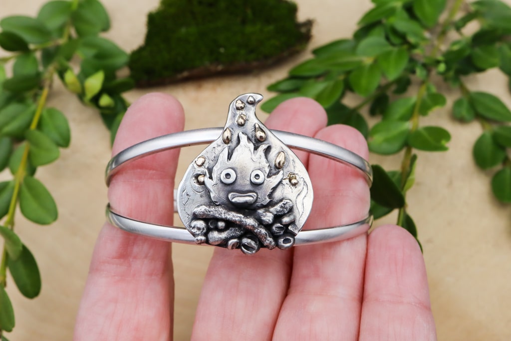 A hand is shown holding the handmade sterling silver Howl's Moving Castle bracelet depicting Calcifer, a fire demon, from the movie.