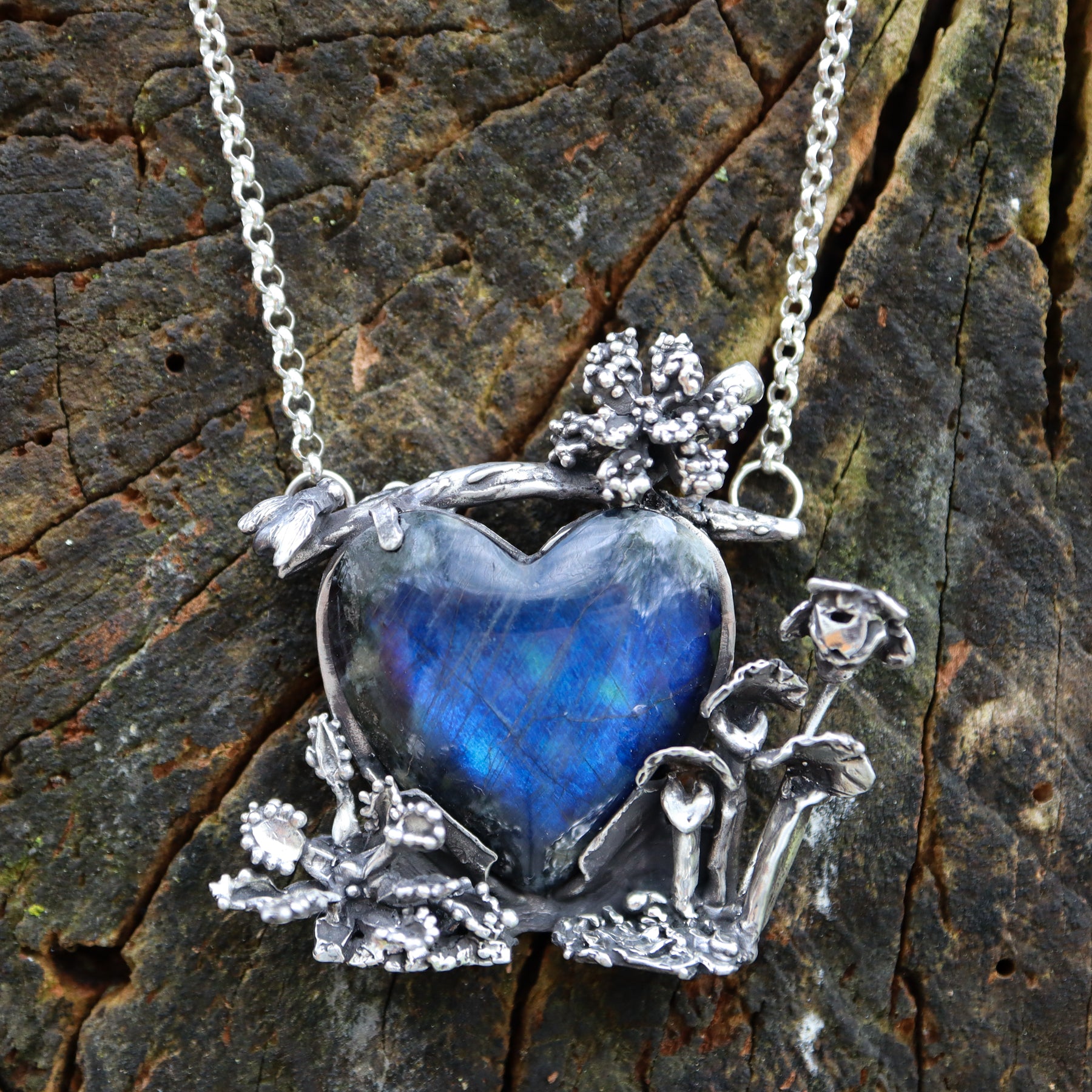 A deep blue spectrolite stone is surrounded by carved silver carnivorous plants with a tiny silver fly on the top left of the pendant. The necklace is shown on a dark brown piece of tree bark.