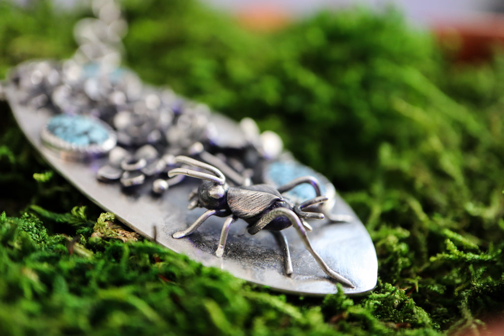 A closer up side view of the hand carved sterling silver cricket. You can see her antennae, legs, wings, and her large eyes. The necklace is sitting in a bed of bright green moss.