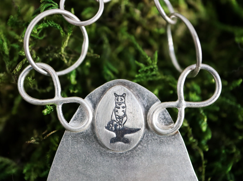 A close up photo of my maker's mark of my striped cat Max sitting on an anvil with his tail curled up around his feet.