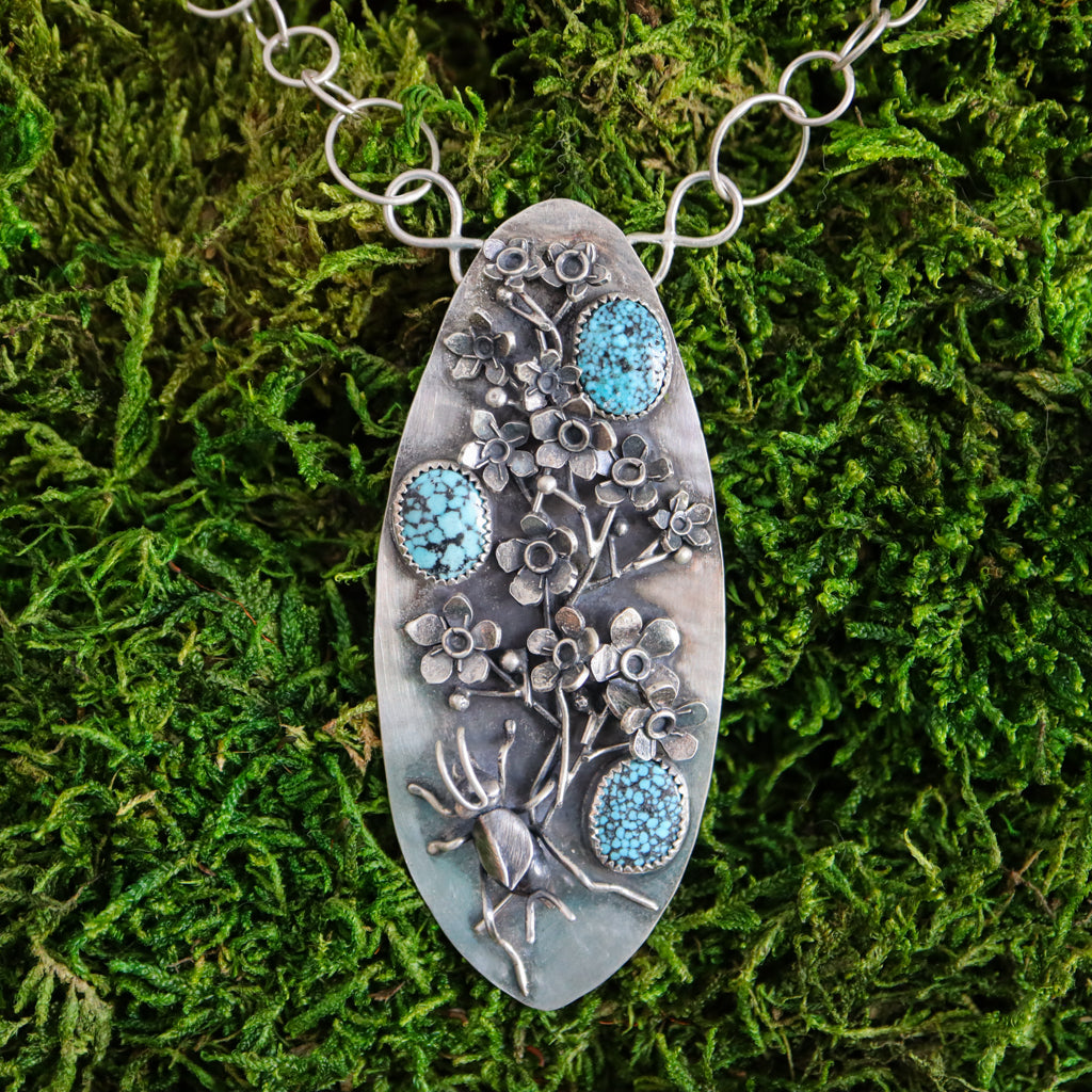 A sterling silver handmade necklace in an elongated shield shape. There is a hand carved cricket at the bottom of the pendant and above her is a sprig of silver forget-me-not flowers. There are three light blue spotted turquoise stones mixed among the flowers. The necklace is shown in a bed of bright green moss. 