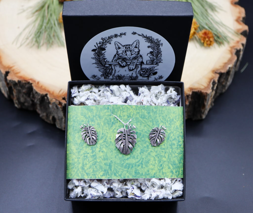A sterling silver monstera deliciosa jewelry set of earrings and a necklace. The set of jewelry is shown in a black jewelry box on top of plantable paper.