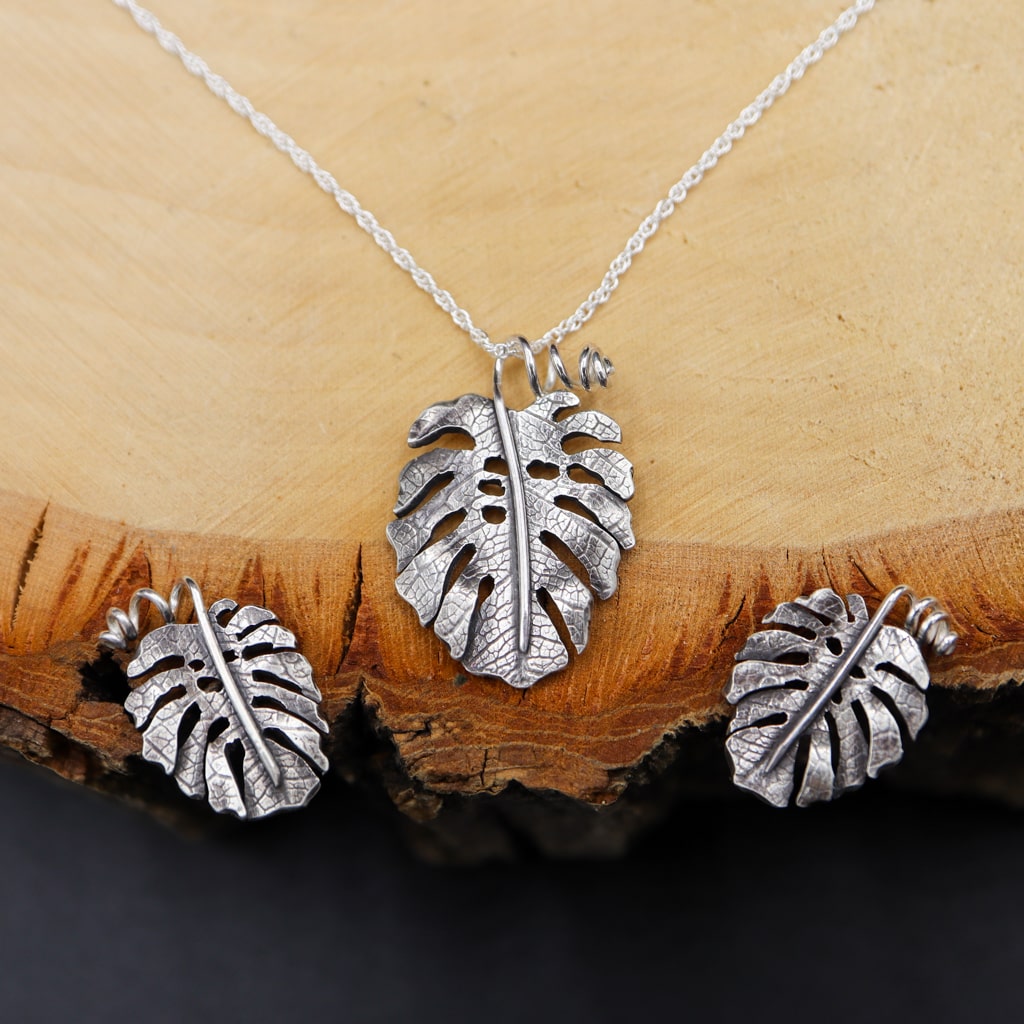 A dainty set of monstera deliciosa stud earrings and a matching necklace. The three pieces of jewelry are shown on a piece of light tan wood.