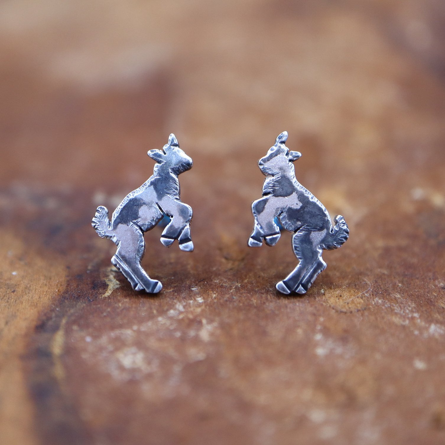 Little handmade sterling silver jumping goat earrings with dark spots and cute fuzzy tails. They are shown on a piece of dark brown wood.