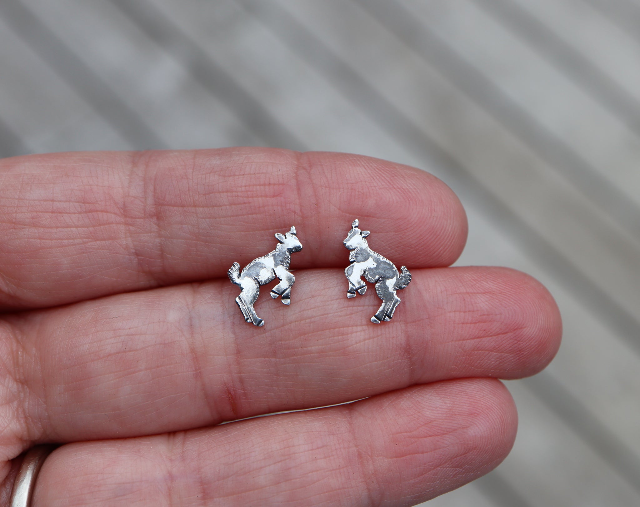 Small silver goat earrings that are kicking up into the air being silly. They have darkened spots on them so that they look black and white. A hand is shown holding them for size reference.