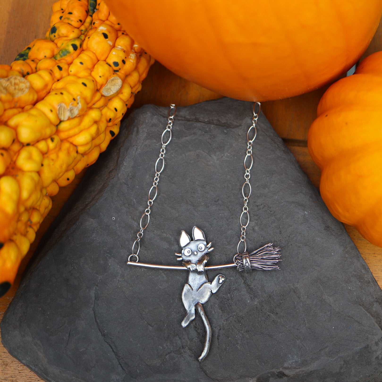 Jiji cat from Kiki's Delivery Service necklace that is handmade from sterling silver. His is trying to hang onto a realistic looking silver broom and attached to a looped chain necklace. The necklace is surrounded by orange pumpkins and gourds and on a dark grey piece of slate.