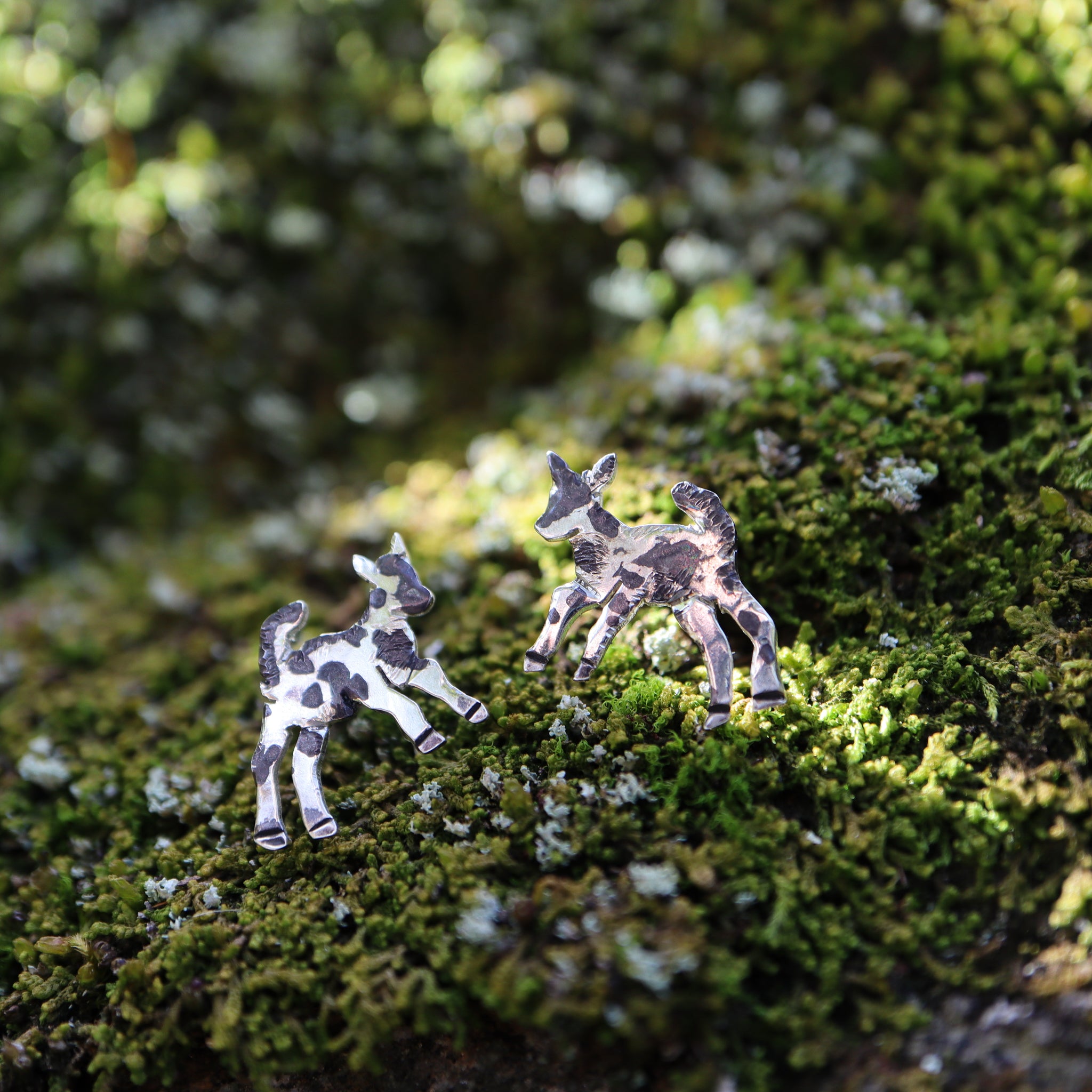 Silly little goat earring studs made from sterling silver and handmade. They are about 1/2 inch wide and given dark spots. The earrings are shown on some live green moss. 