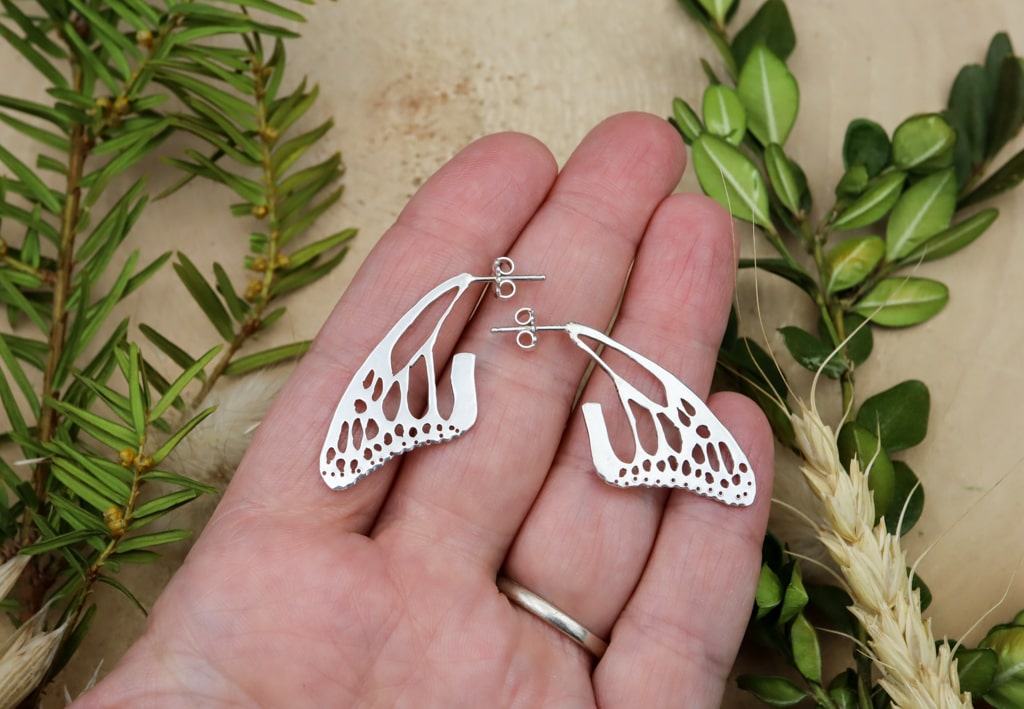 A hand is shown holding the handmade sterling silver monarch wing stud earrings. They are about 1.25 inches wide.