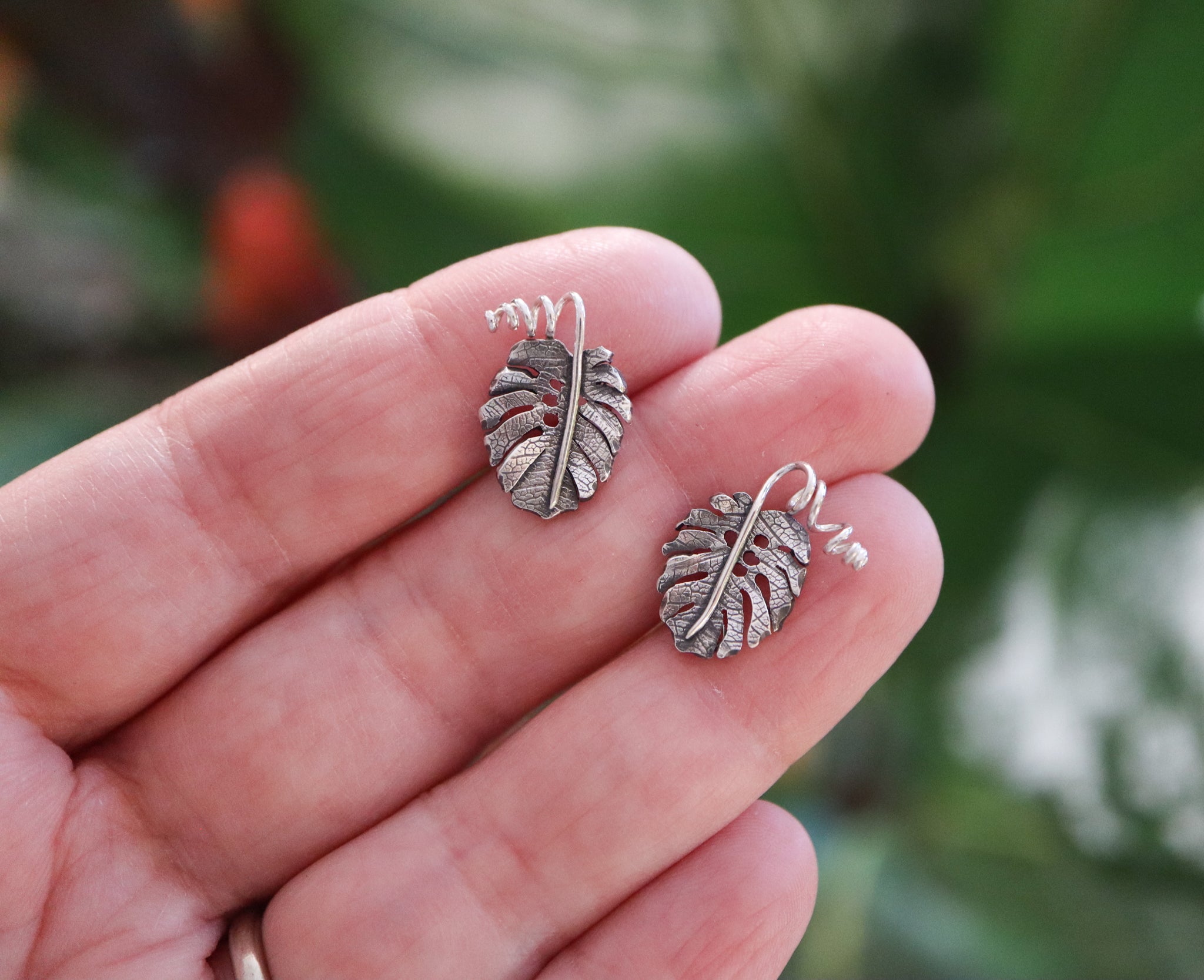 A hand is shown holding a pair of sterling silver monstera deliciosa earring studs. They earring are about 1/2 inch tall and have little twirled stems.