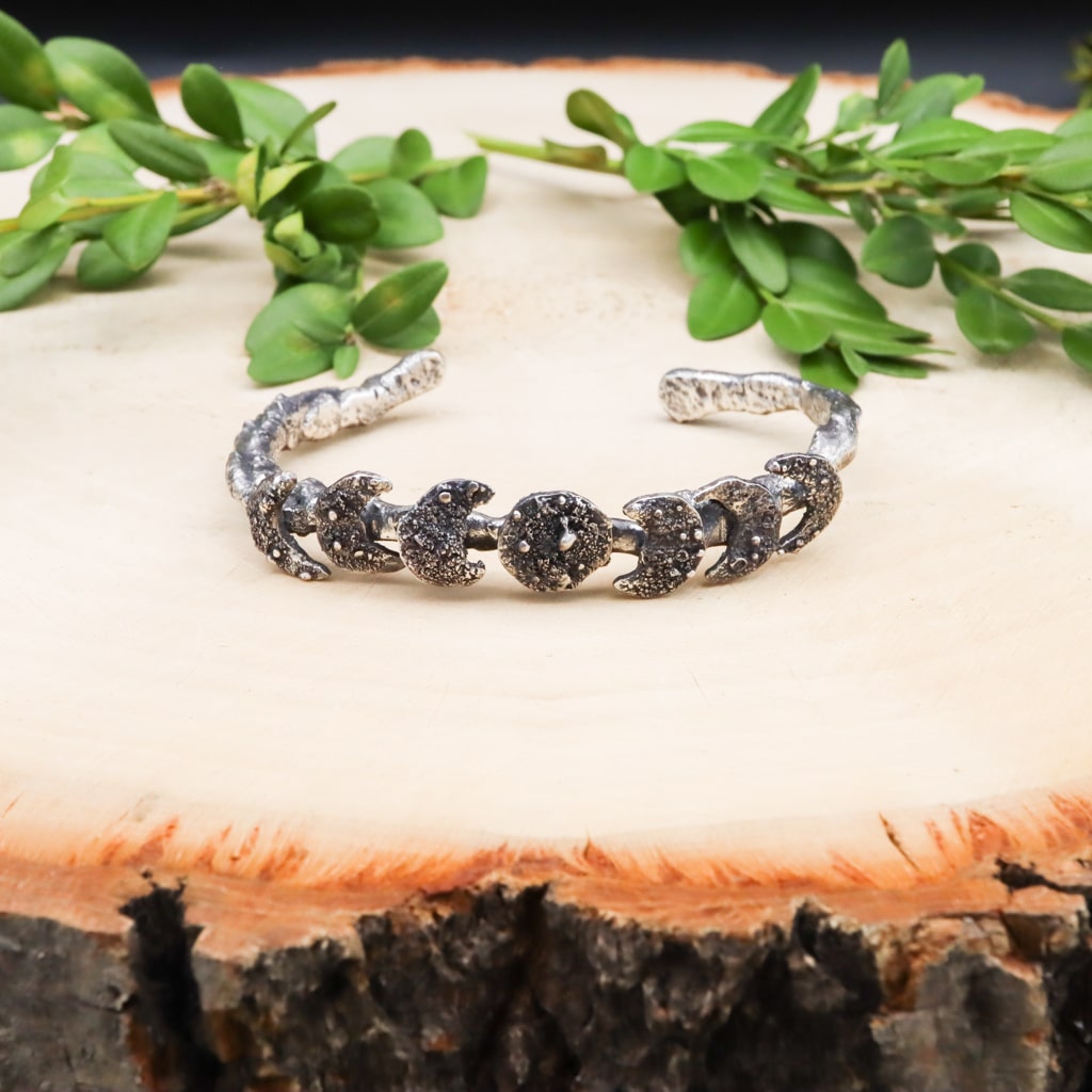 A sterling silver hand carved moon phase bracelet. There are 7 different moons on the bracelet next to each other in a line. The bracelet band looks like a curved tree branch. The bracelet is shown on a piece of light tan wood with greenery behind it.