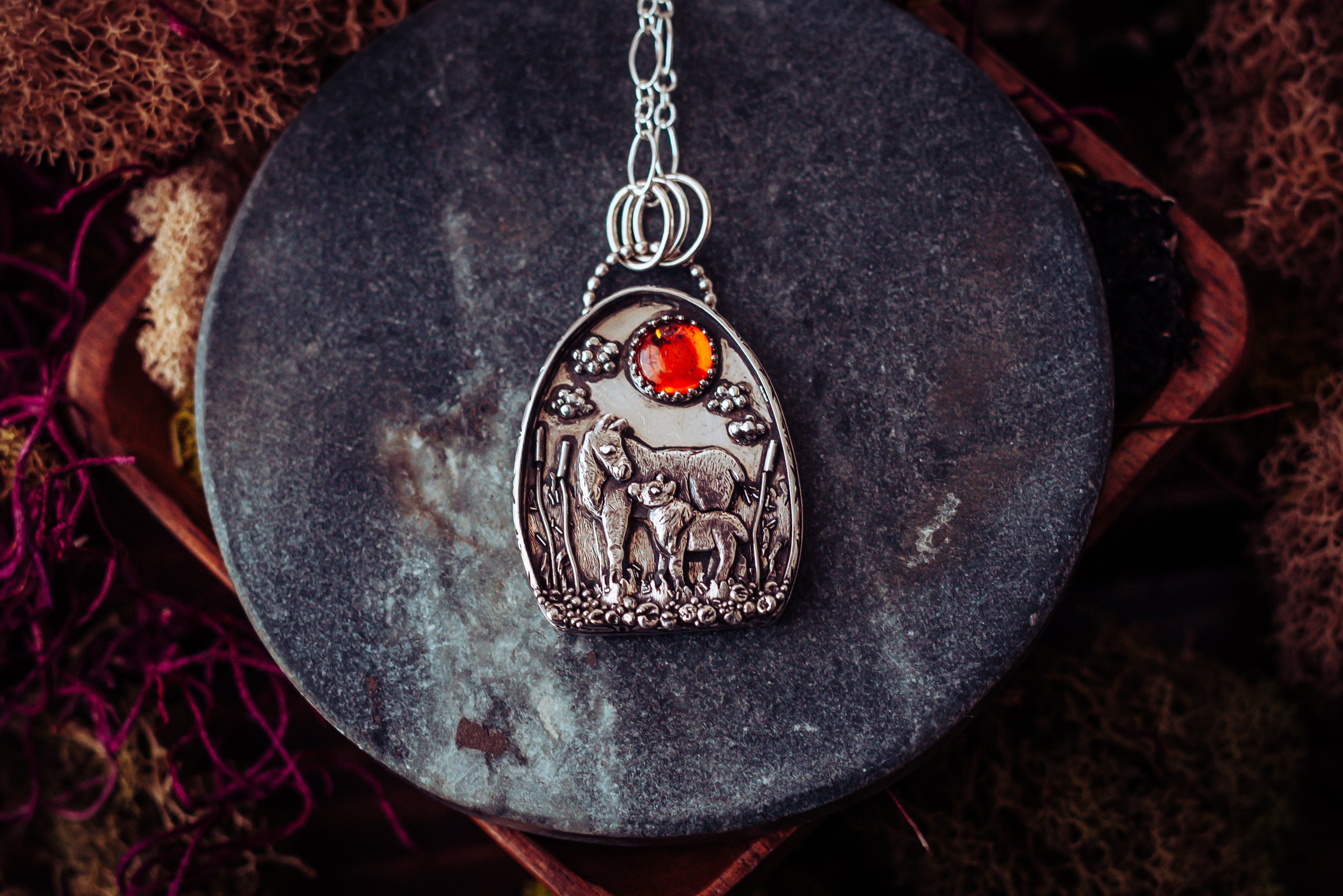 A mother and baby moose pendant made from sterling silver. It has a deep orange round amber stone at the top to represent the sun and they are surrounded by cat tails, stones, and plants. The pendant is on a dark grey and while stone and surrounded by mosses.