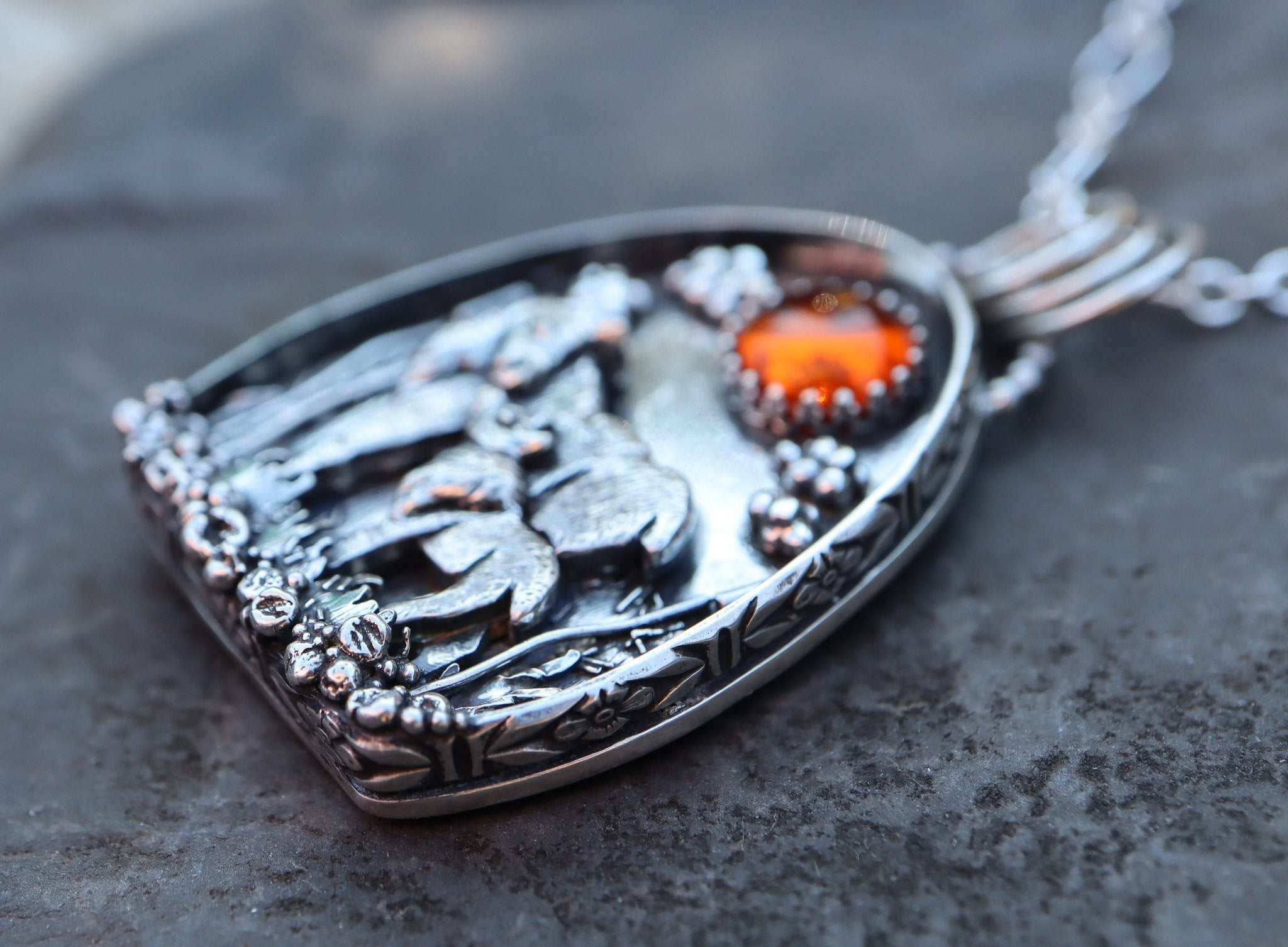 A side view of the shadowbox moose pendant shown with an amber stone on a slate colored piece of stone. Made by The Striped Cat Metalworks.