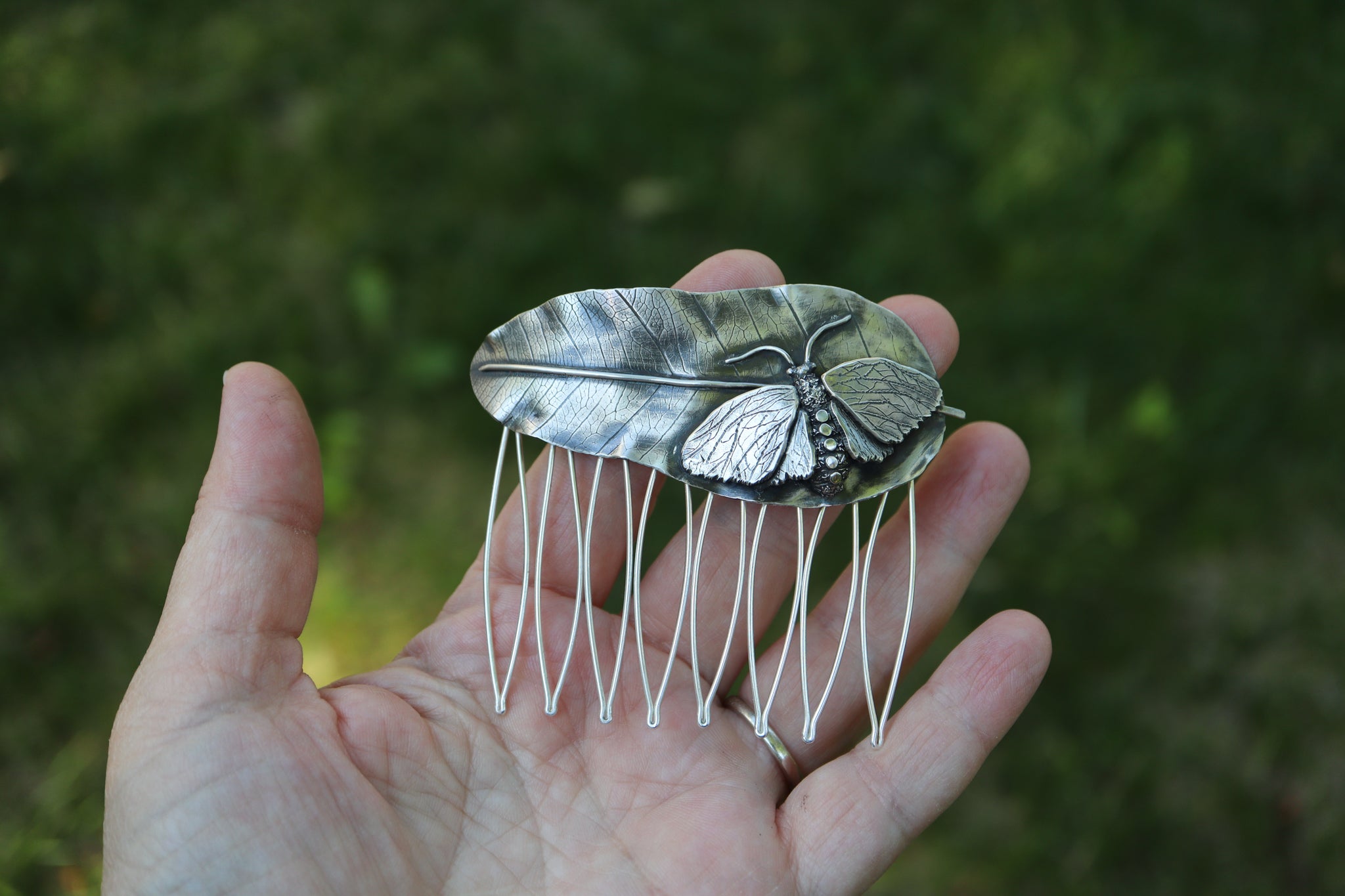 A hand is shown holding the milkweed tussock moth hair comb to show size. It is about 3.25 inches tall and wide.