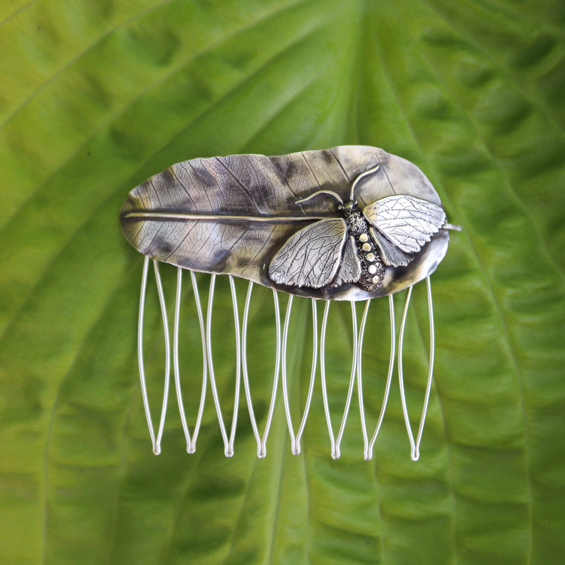 A sterling silver hair comb depicting a hand carved milkweed tussock moth on one end of the top, sits on top of a silver milkweed leaf. The hair comb is about 3.25 inches tall and wide. The hair comb is show on top of a light green plant leaf.