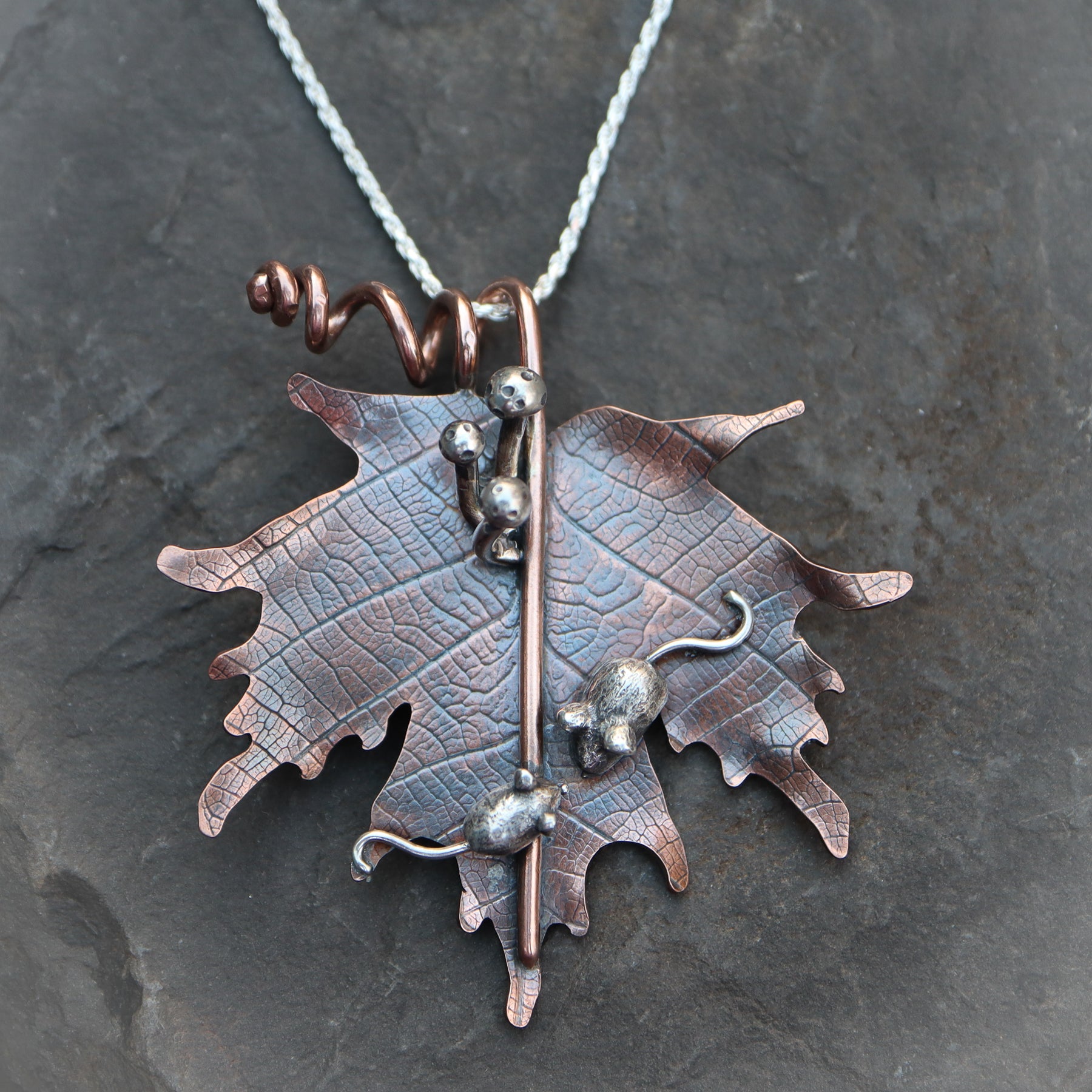 A copper maple leaf that is about 2 inches tall and 2 inches wide. On the leaf are two hand carved sterling silver mice, a mother and a baby, snuggled next to each other. Above the mice are three silver mushrooms coming from the stem of the maple leaf. The necklace is shown in a dark grey piece of slate.
