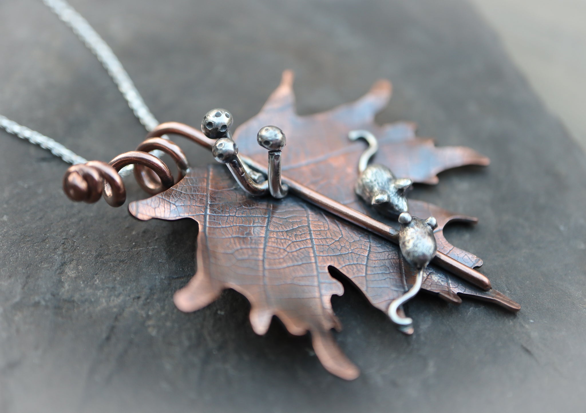 A close up of the three silver mushrooms on the copper maple leaf. The leaf is shown on a piece of dark grey stone slate.