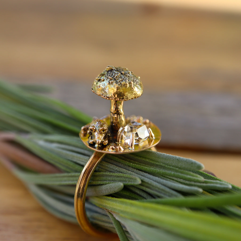 A 14 karat gold ring that features a tiny hand carved mouse sitting under a mushroom. Next to them is a shiny herkimer diamond point that is prong set. The ring is shown on a sprig of lavender. 