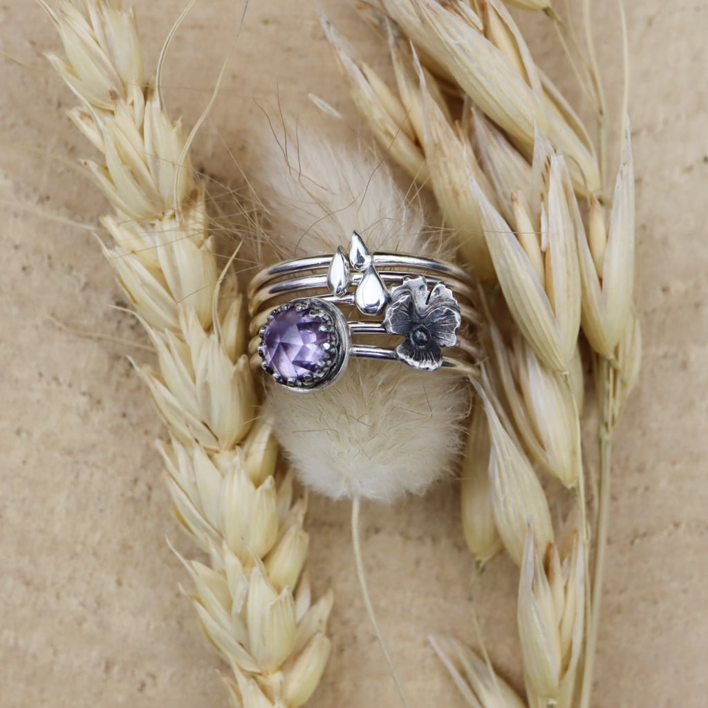 A set of 5 sterling silver stacking rings. There are three rings that each have a tiny carved raindrop on them. One band has a miniature pansy flower. One band has a light purple rose cut amethyst gemstone. They are shown on a fluffy piece of grass next to wheat heads.