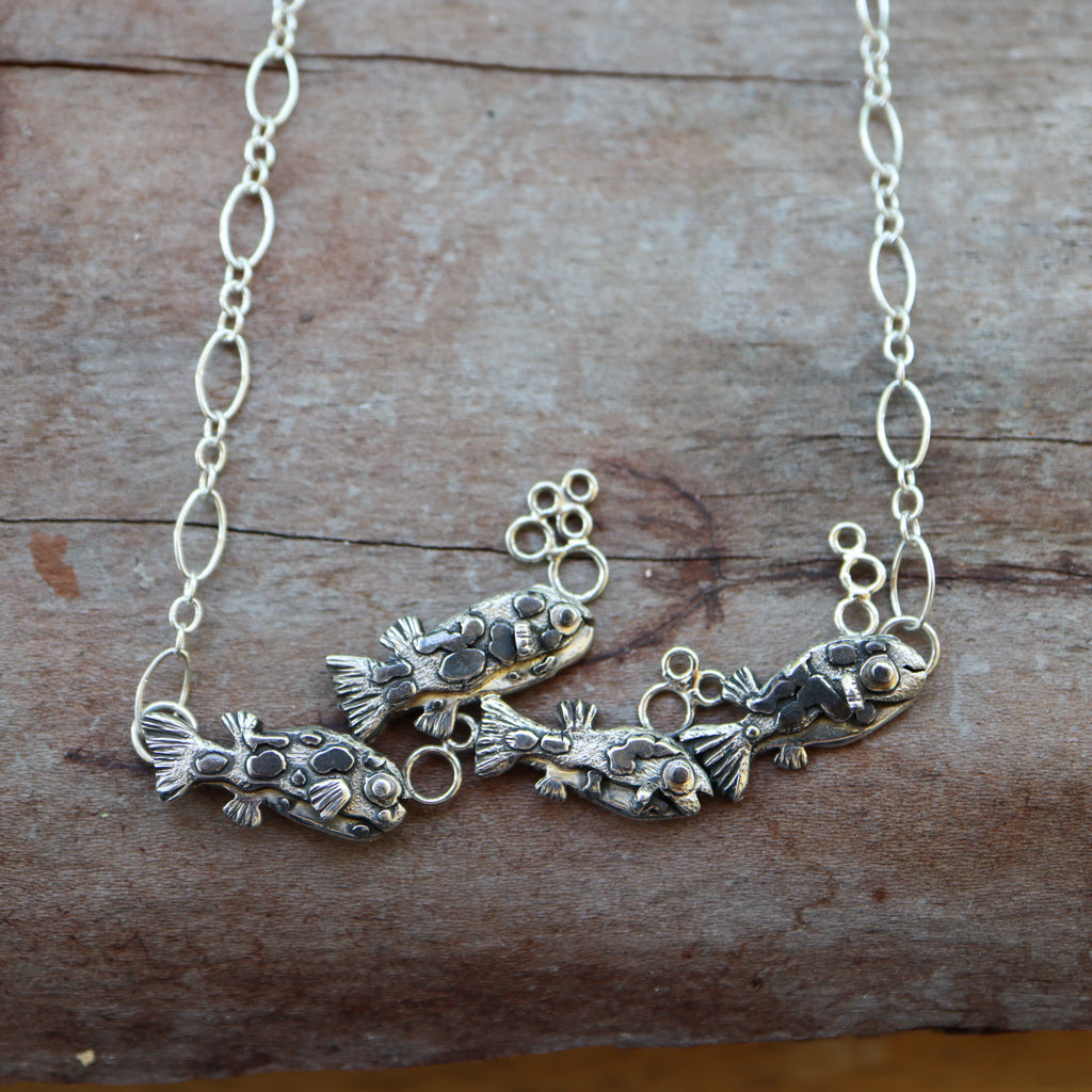 A sterling silver dwarf pea puffer necklace featuring 4 little fish swimming in a row behind each other. All of the fish have a few silver bubbles above them. They are on a silver looped necklace and shown on a piece of dark wood.
