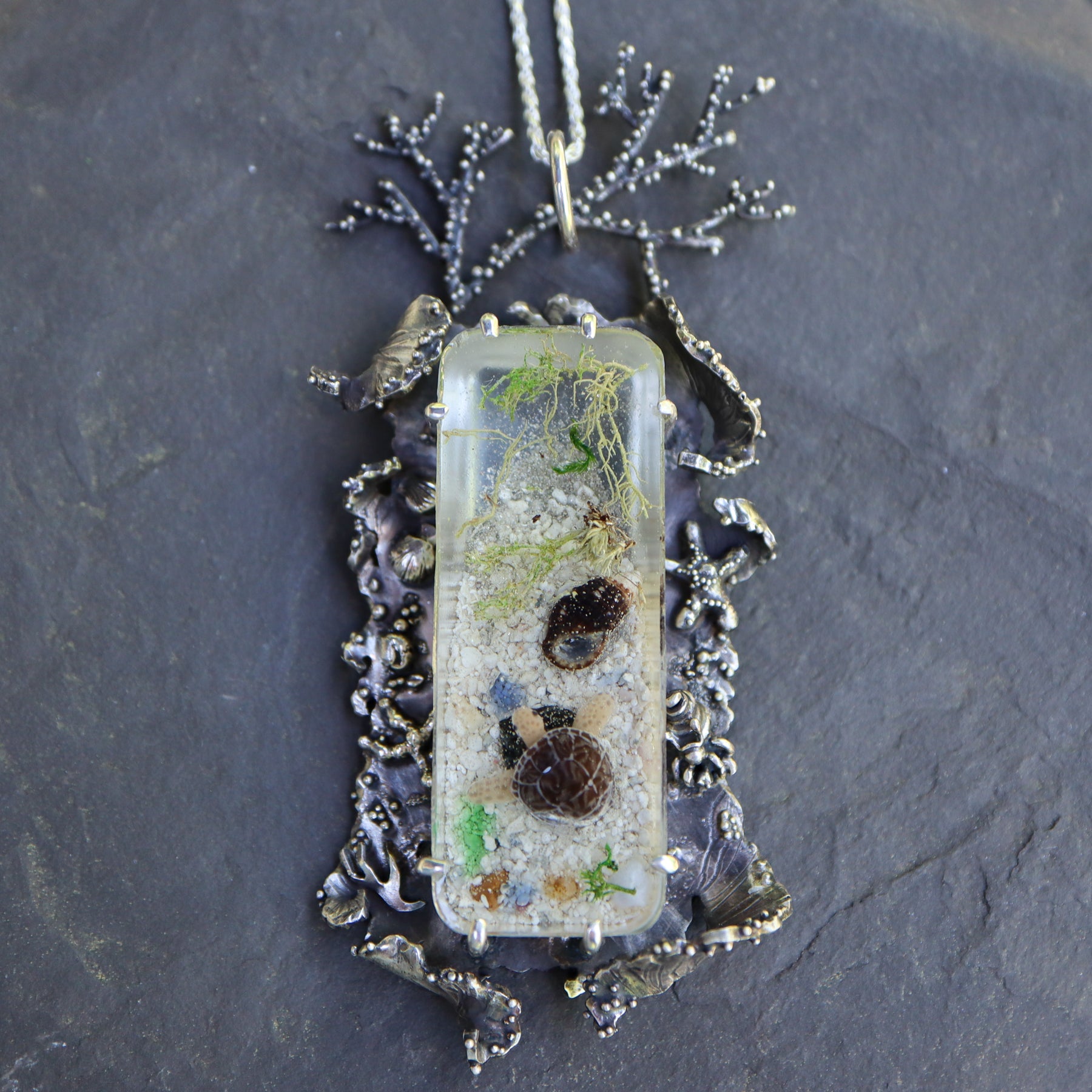 A handmade glass sea turtle baby is set inside a resin cabochon with different colors of sea glass shards, seaweed, and sand. The cabochon is held in place with prngs in the center of the sterling silver pendant. The pedant is made to look like rolling waves on all four sides and has starfish, hermit crabs, seaweed, and seashells. The pendant is about 3 inches tall and 1.5 inches wide and shown on a dark grey piece of stone.