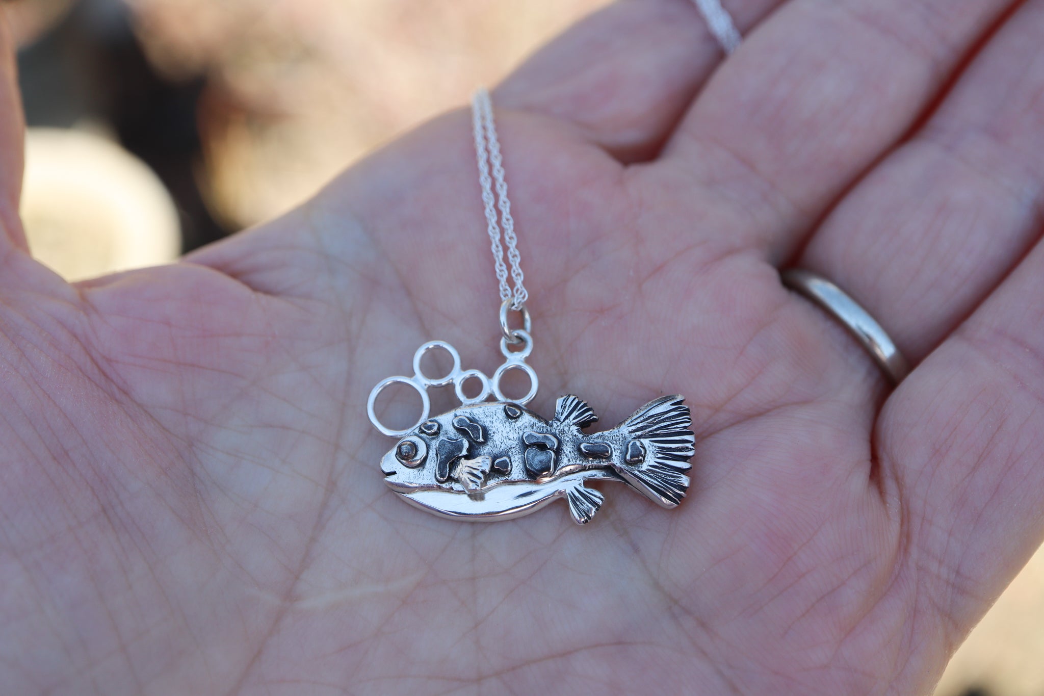A hand is shown holding a 1 inch dwarf pea puffer necklace for size scale.