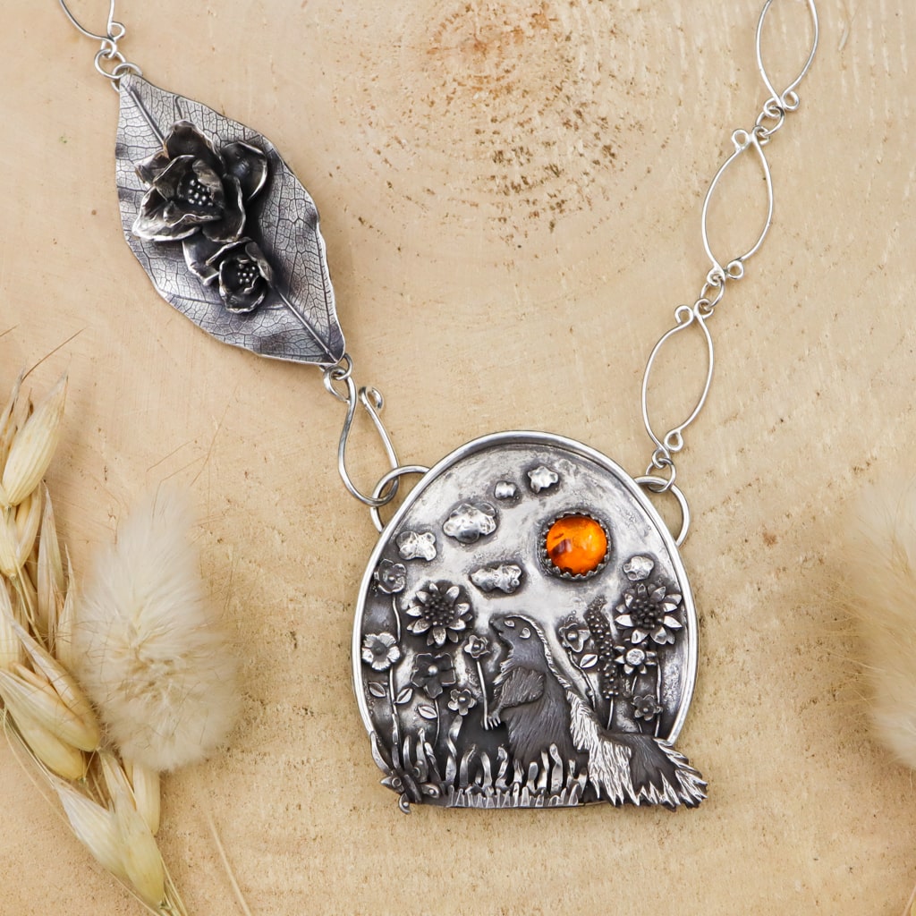 A handmade sterling silver skunk necklace. She is surrounded by lots of silver flowers of all kinds. Above her in the sky are silver clouds and a small, round, amber cabochon to represent the sun. Along the necklace chain is a handmade cluster of flowers. The necklace is shown on a light tan piece of wood and fluffy grasses.