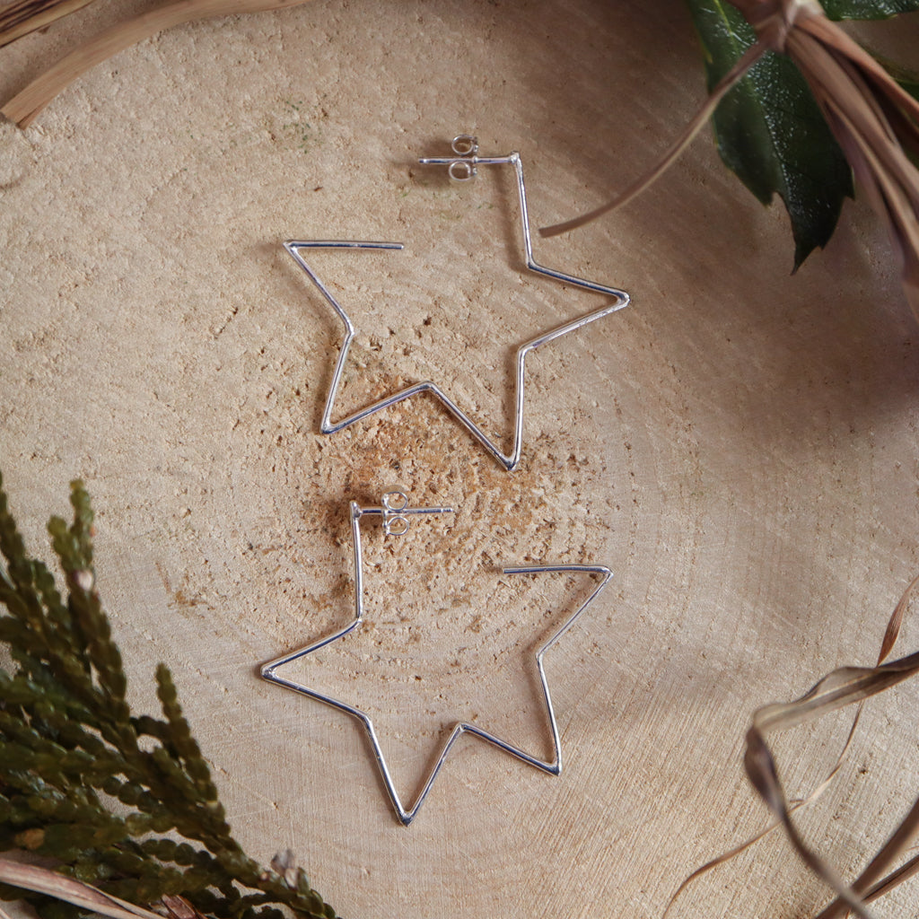 Sterling silver handmade star hoop earrings are shown on a piece of light tan wood.