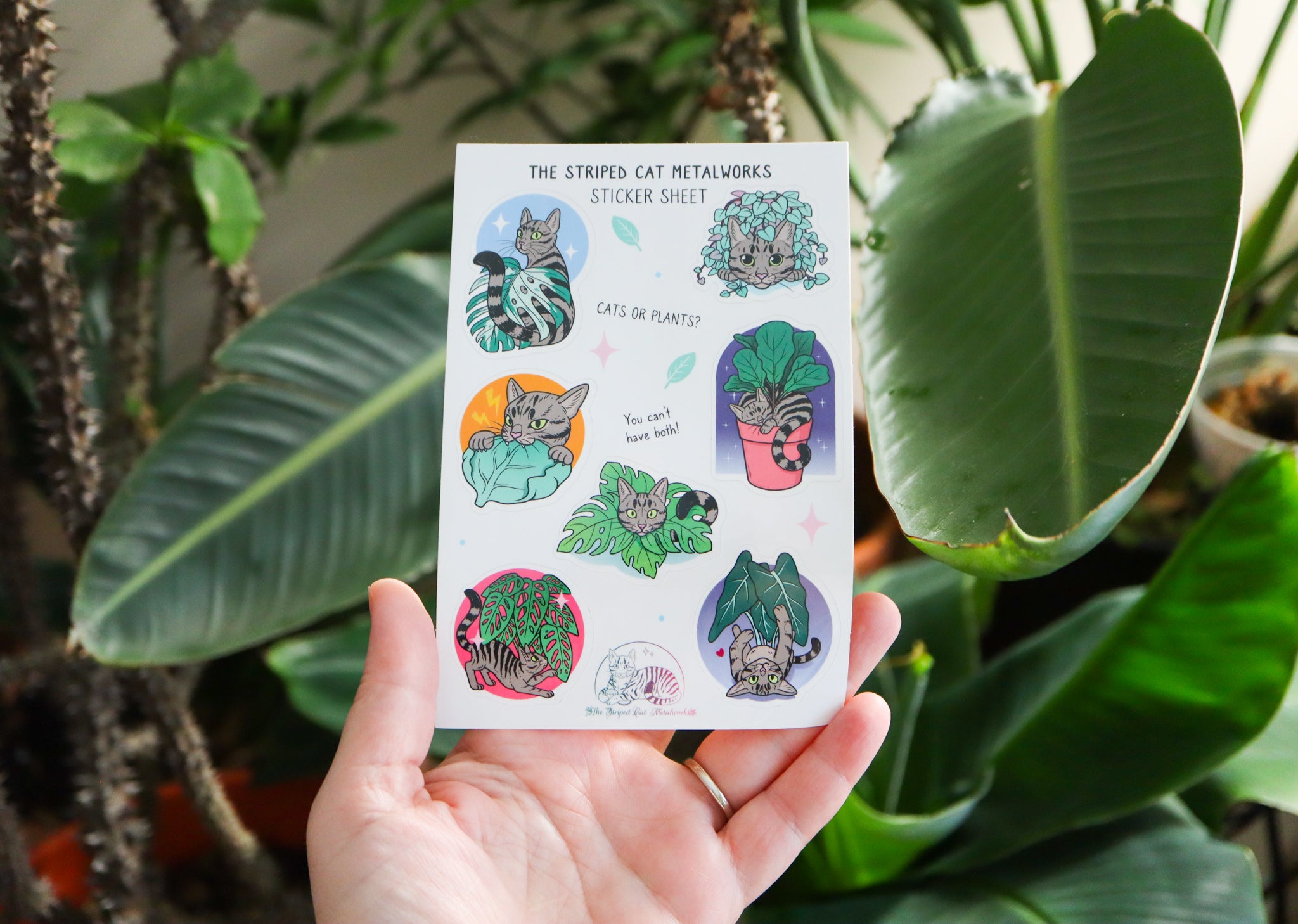 The Striped Cat Metalworks sticker sheet featring MAx the striped cat and houseplants like a fiddle leaf fig, monstera adansonii, monstera albo, string of hearts, and an alocasia polly all being played with by Max the cat! A hand is holding the sticker sheet in front of houseplants to show the size.