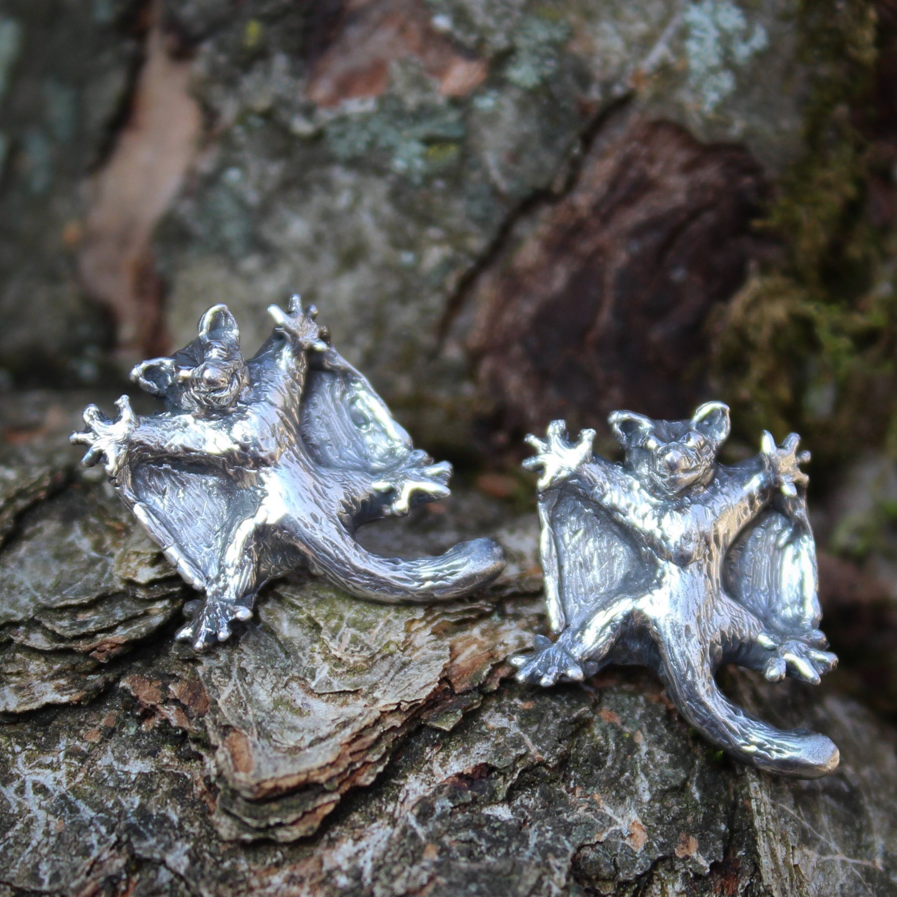 Handmade sterling silver sugar glider earrings. They are about 1/2 inch tall and shown on a piece of tree bark.
