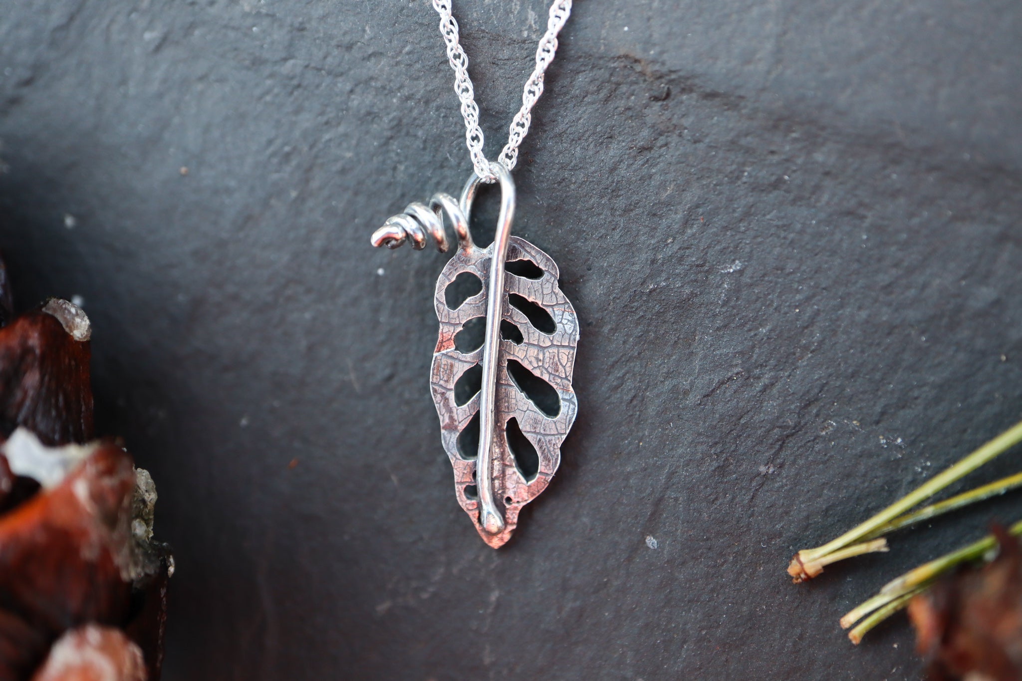 A small silver monstera leaf necklace is shown on a piece of dark grey slate. 