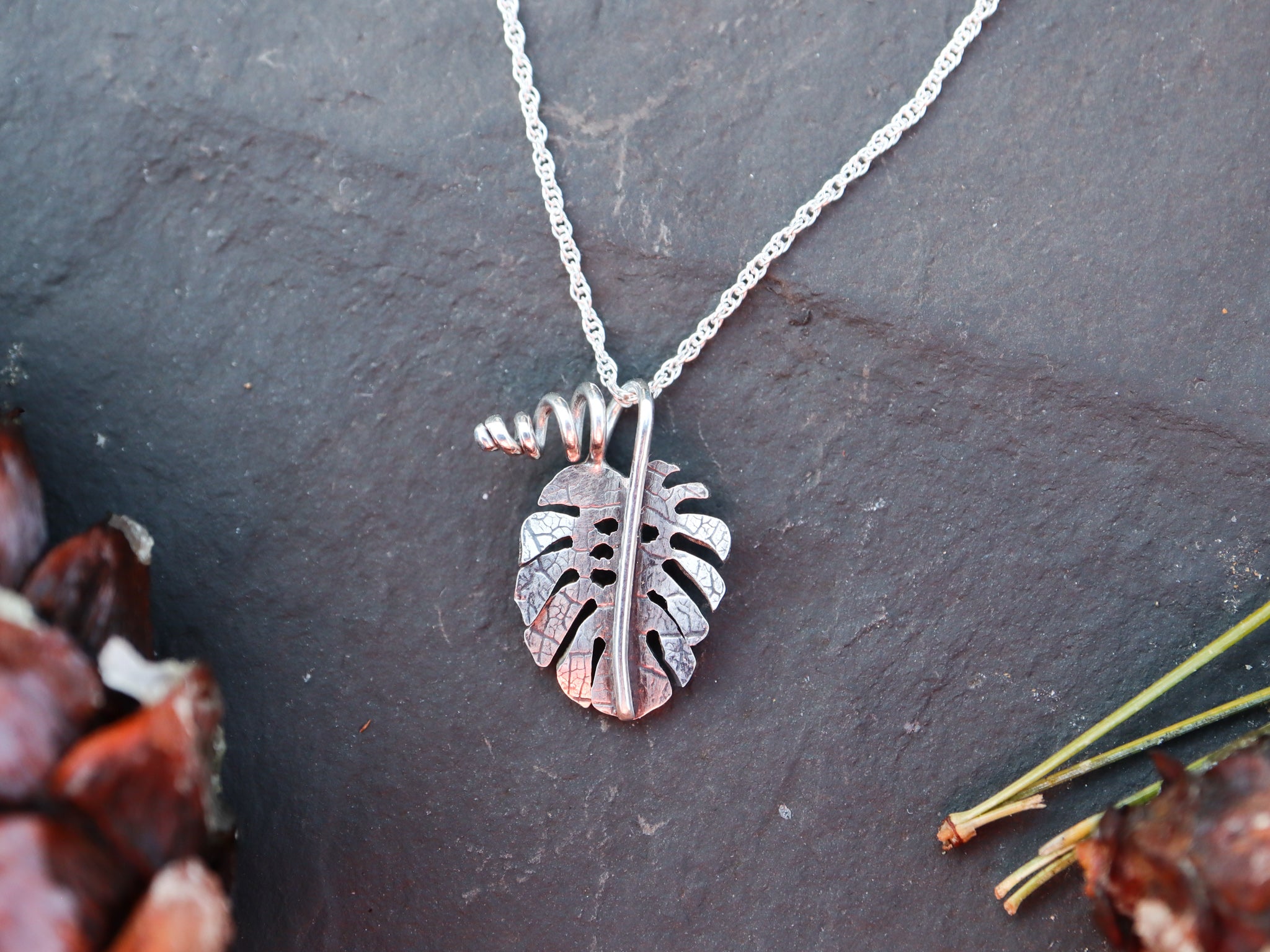 A small sterling silver monstera deliciosa pendant shown in front of a dark gray slate tile