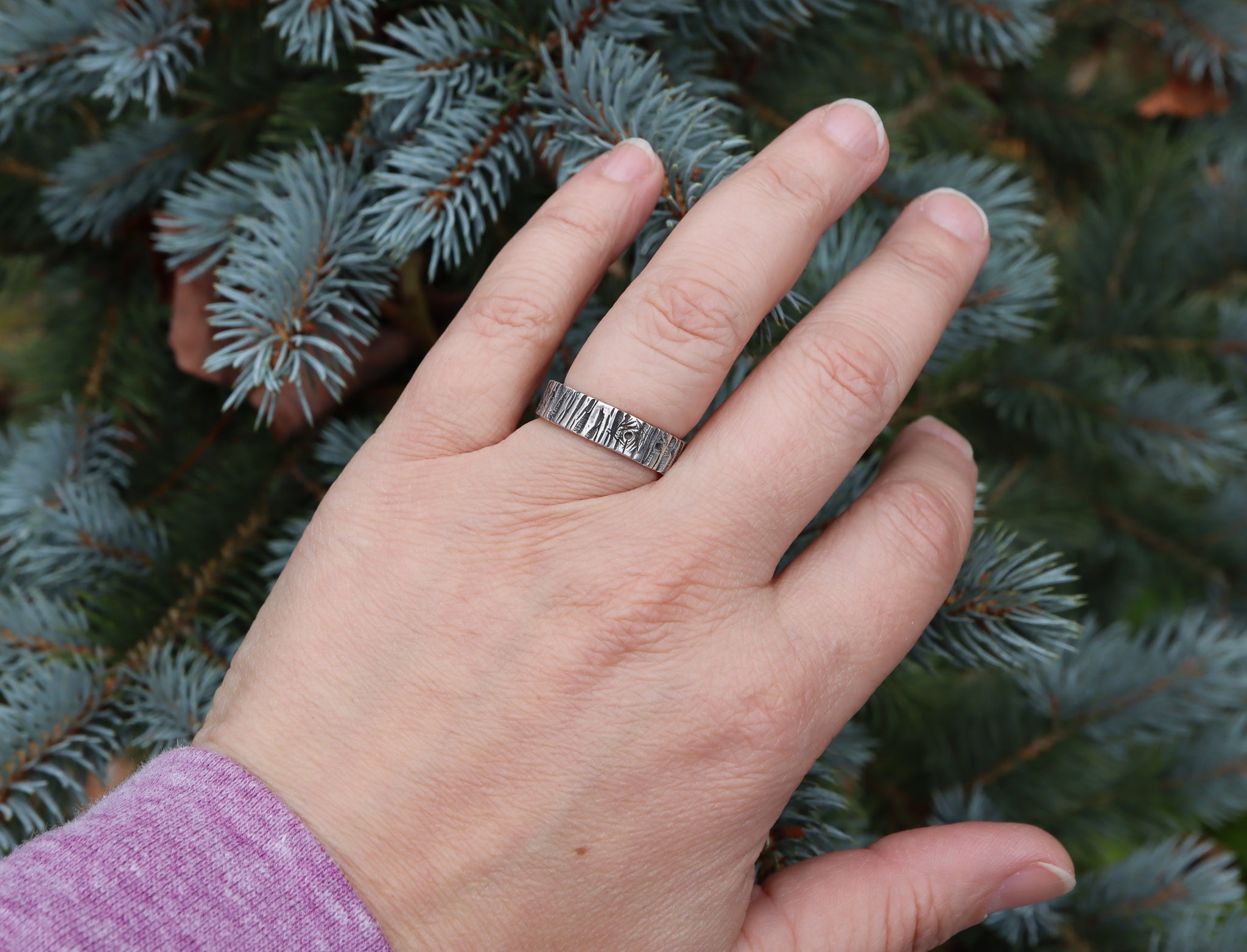 Someone wearing a sterling silver handmade tree bark ring pictured in front of an evergreen tree. The ring is made by The Striped Cat Metalworks in Massachusetts.