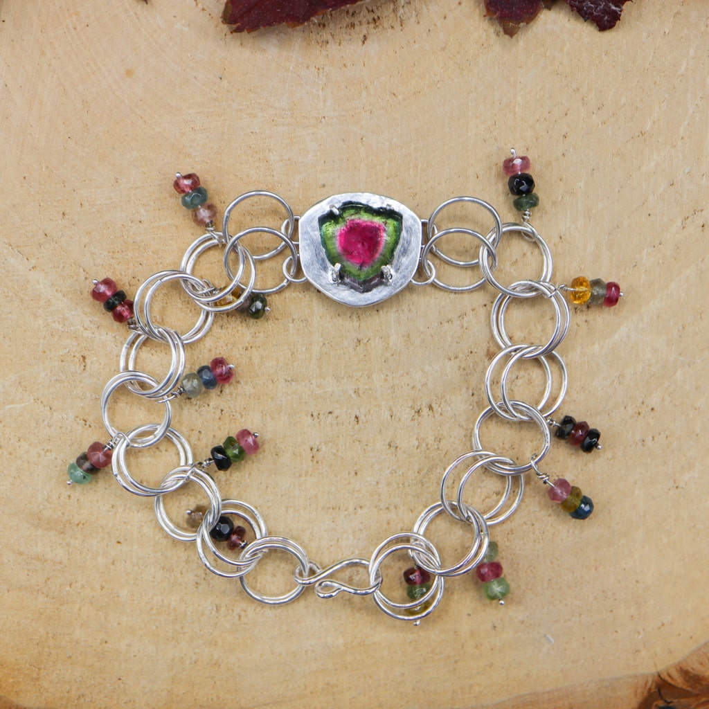 Watermelon tourmaline bracelet with a central pink and green roughly round shape stone. There are large circular links that make up the bracelet. On each link there is a little strand of three multi colored tourmaline stones that move around when the bracelet is being worn. It is shown on a piece of light tan wood.