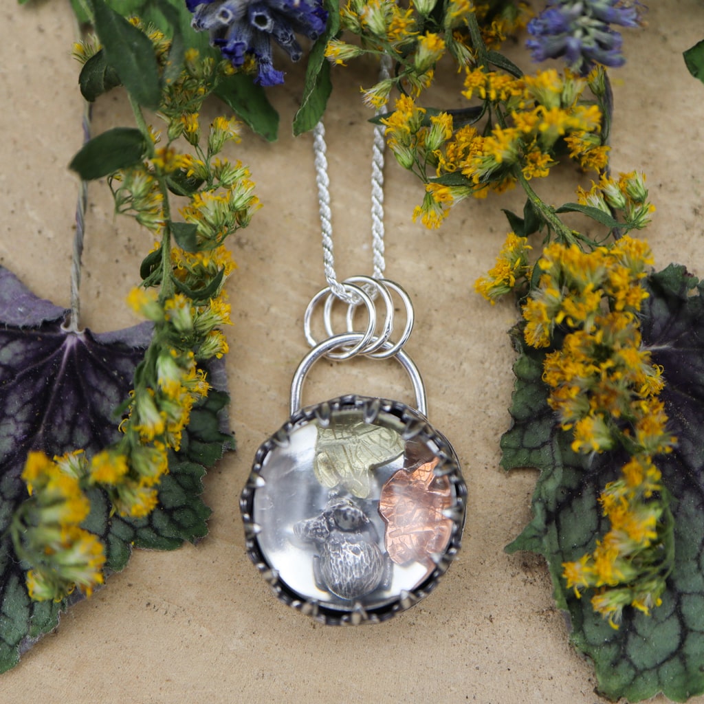 A sterling silver pendant featuring a clear quartz cabochon on top so that you can see a tiny silver mouse and three fall leaves inside. There are purple lavender flower and yellow goldenrod next to the necklace. 